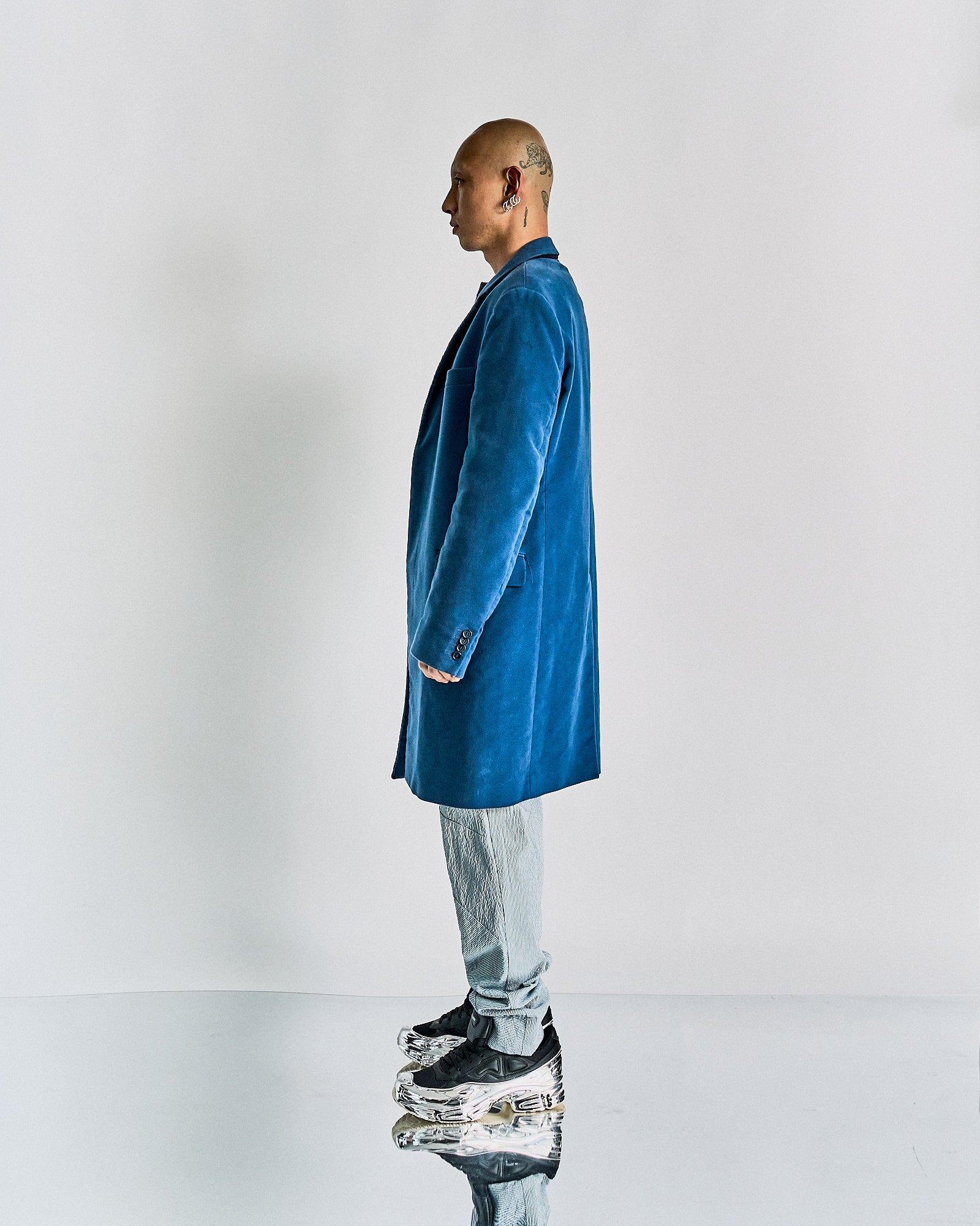 Profile of a bald person with tattoos, wearing a blue velvet Helmut Lang coat and light pants. They stand on reflective shoes, against a plain background. Modern and stylish.