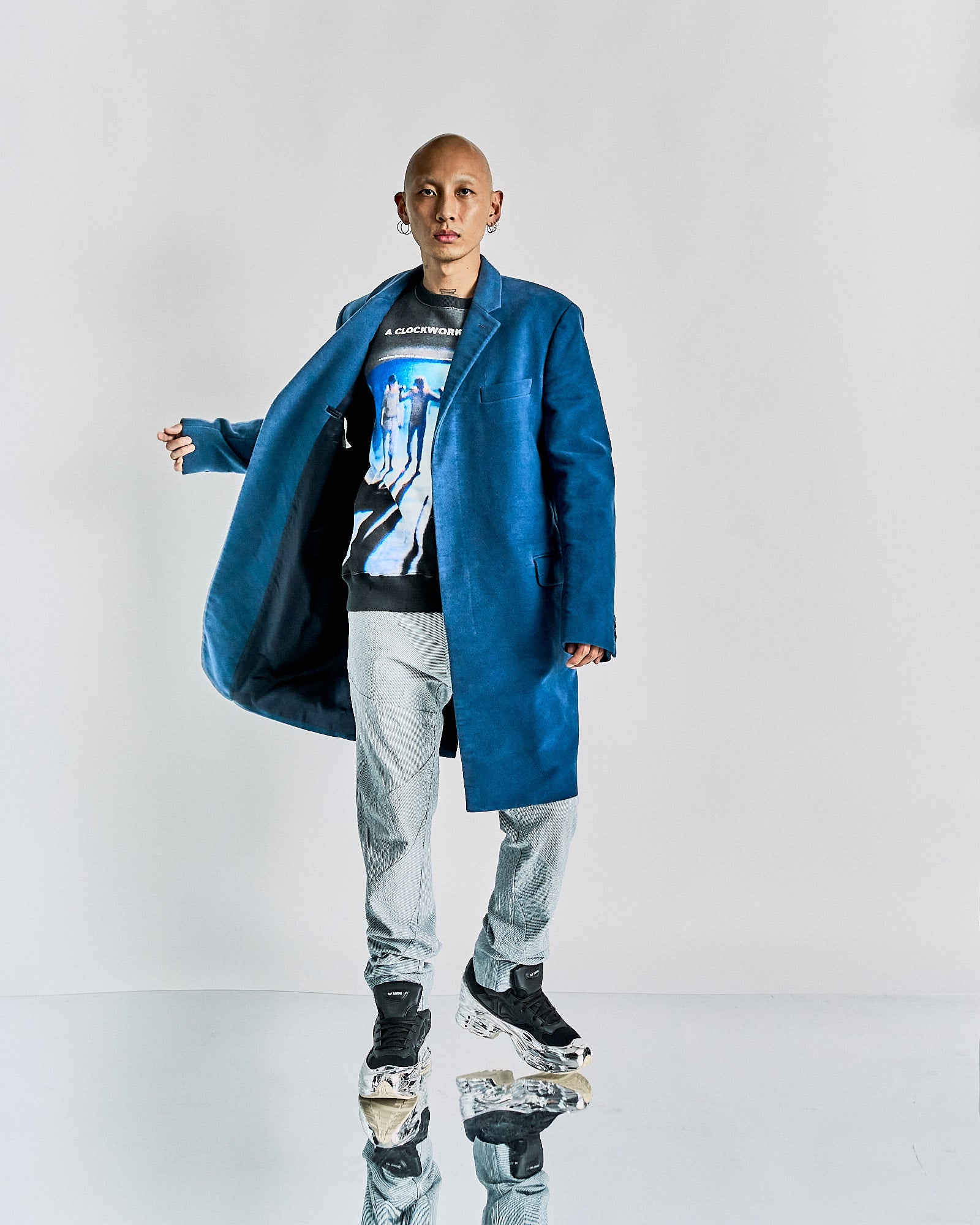 Person wearing a bold blue Helmut Lang coat, graphic T-shirt, and patterned pants with black sneakers. Stands confidently on a reflective surface. Modern and stylish.