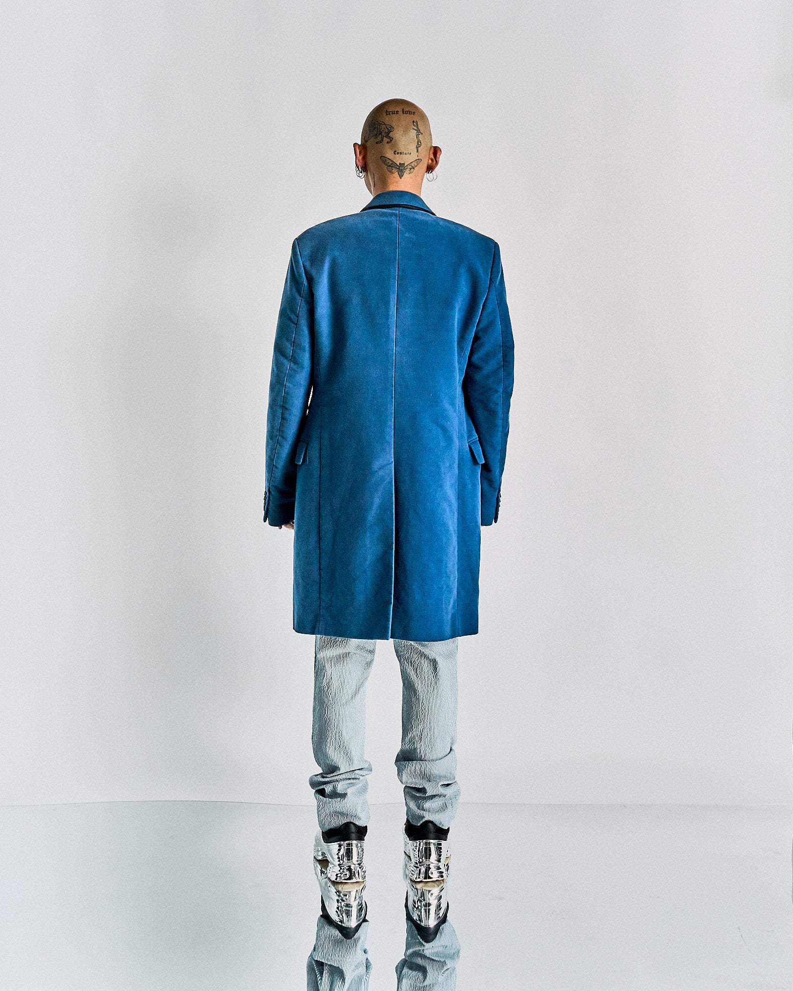 Bald person facing away, showcasing neck tattoos, wears a blue Helmut Lang coat over striped pants. Reflective silver boots suggest a bold, futuristic style.