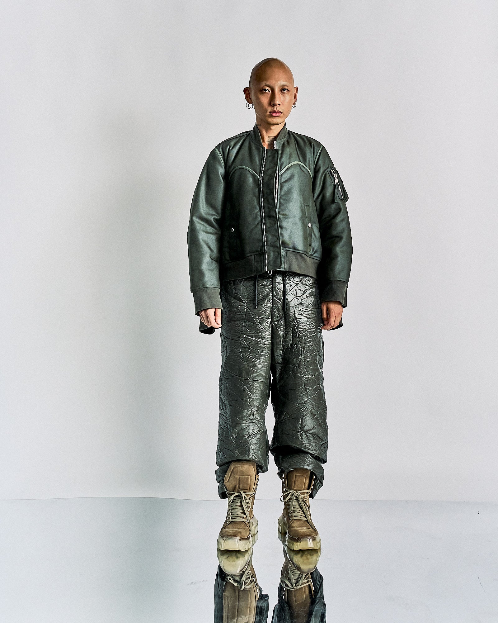 Bald individual in a green Helmut Lang bomber jacket and textured dark pants, paired with tan boots, stands on a reflective floor against a plain backdrop, exuding confidence.