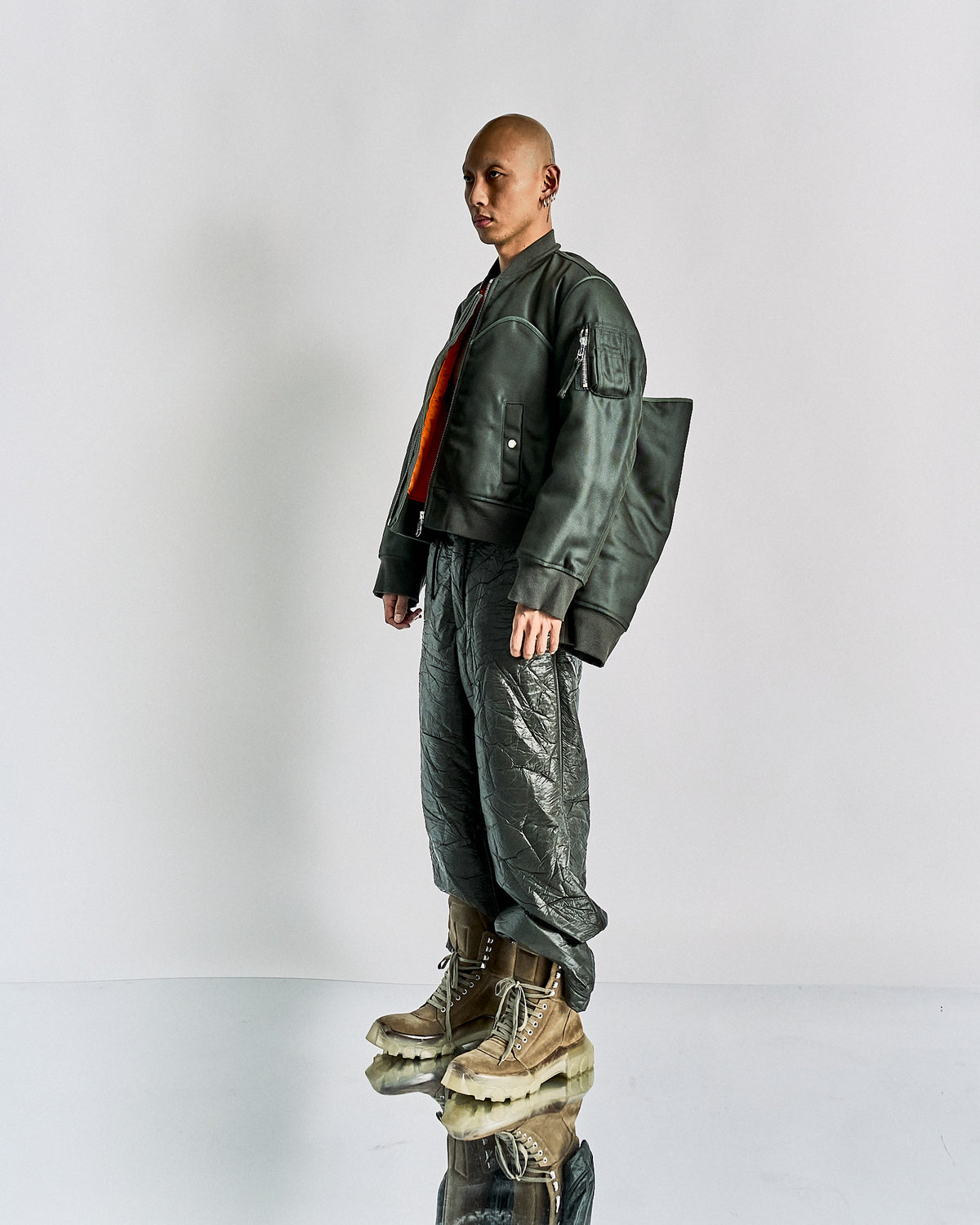 Person in an oversized green Helmut Lang jacket and shiny textured pants bends forward, appearing off-balance. They wear large brown boots. The setting is minimalistic.