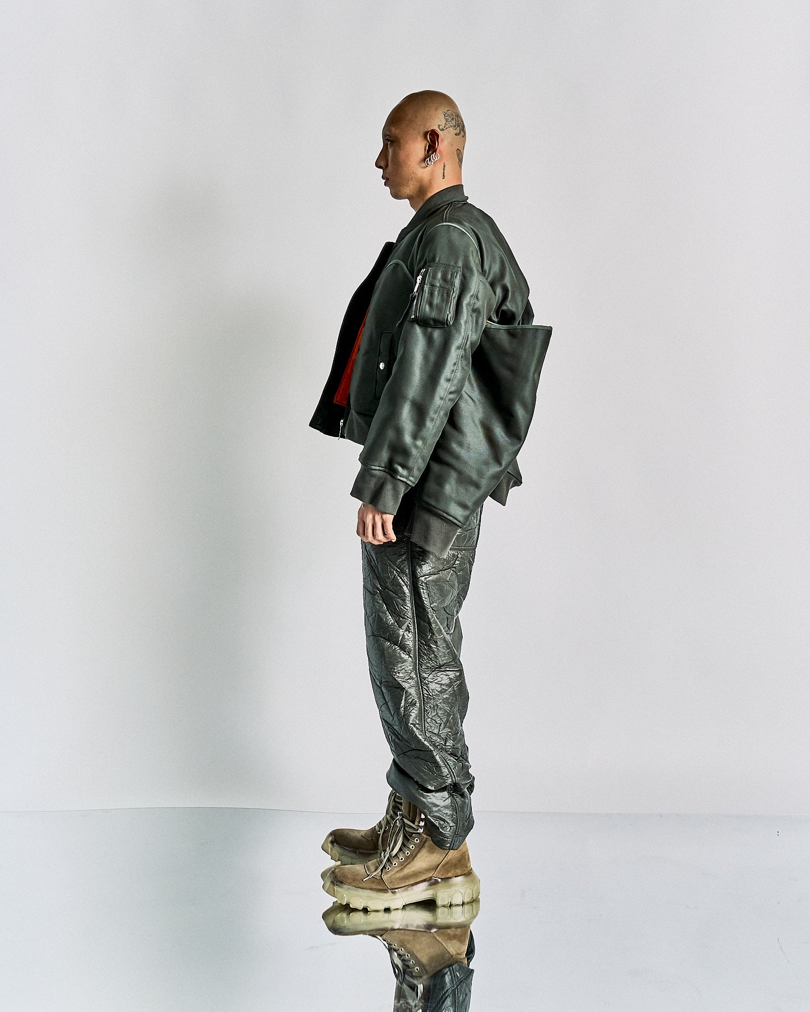 A person stands in profile wearing a green Helmut Lang bomber jacket, quilted pants, and tan boots with thick soles. The look is edgy and futuristic.