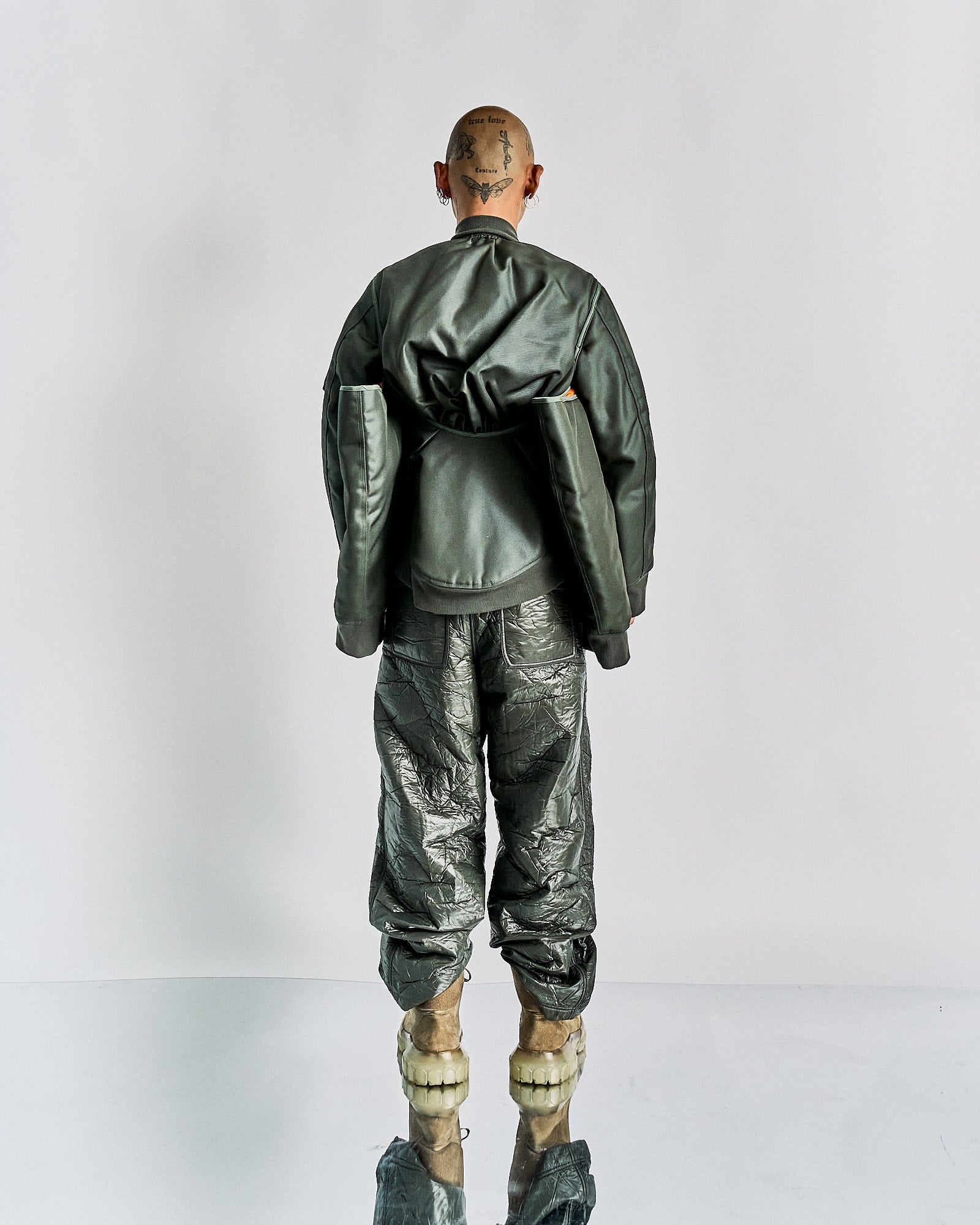 A person stands facing away in futuristic dark green clothing, with a Helmut Lang bomber jacket and crinkled pants. Their bald head is tattooed, and shoes are avant-garde.