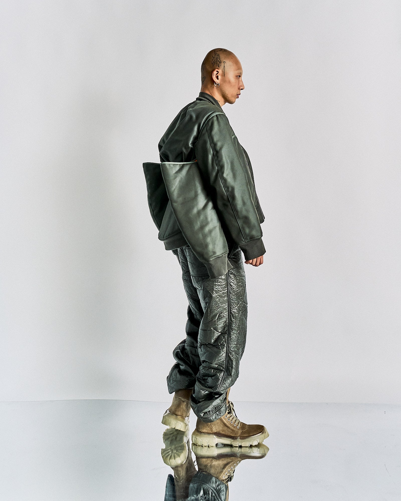 A person stands in profile against a plain background wearing an oversized, dark green Helmut Lang bomber jacket, shiny textured pants, and tan boots with thick soles. The style is modern and edgy.