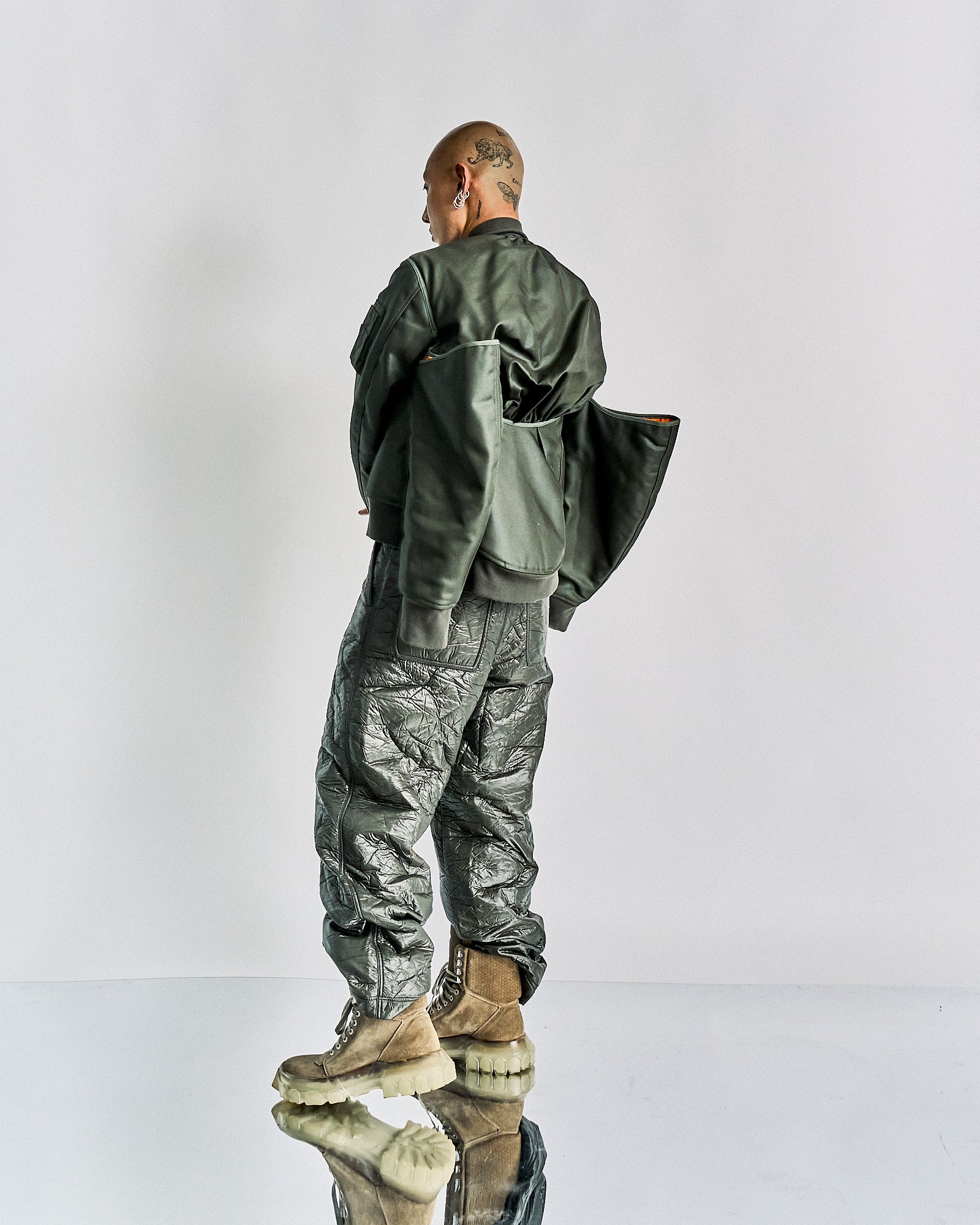 Bald person with tattoos, wearing an oversized green Helmut Lang bomber jacket and textured pants, stands on a reflective surface. The tone is edgy and avant-garde.