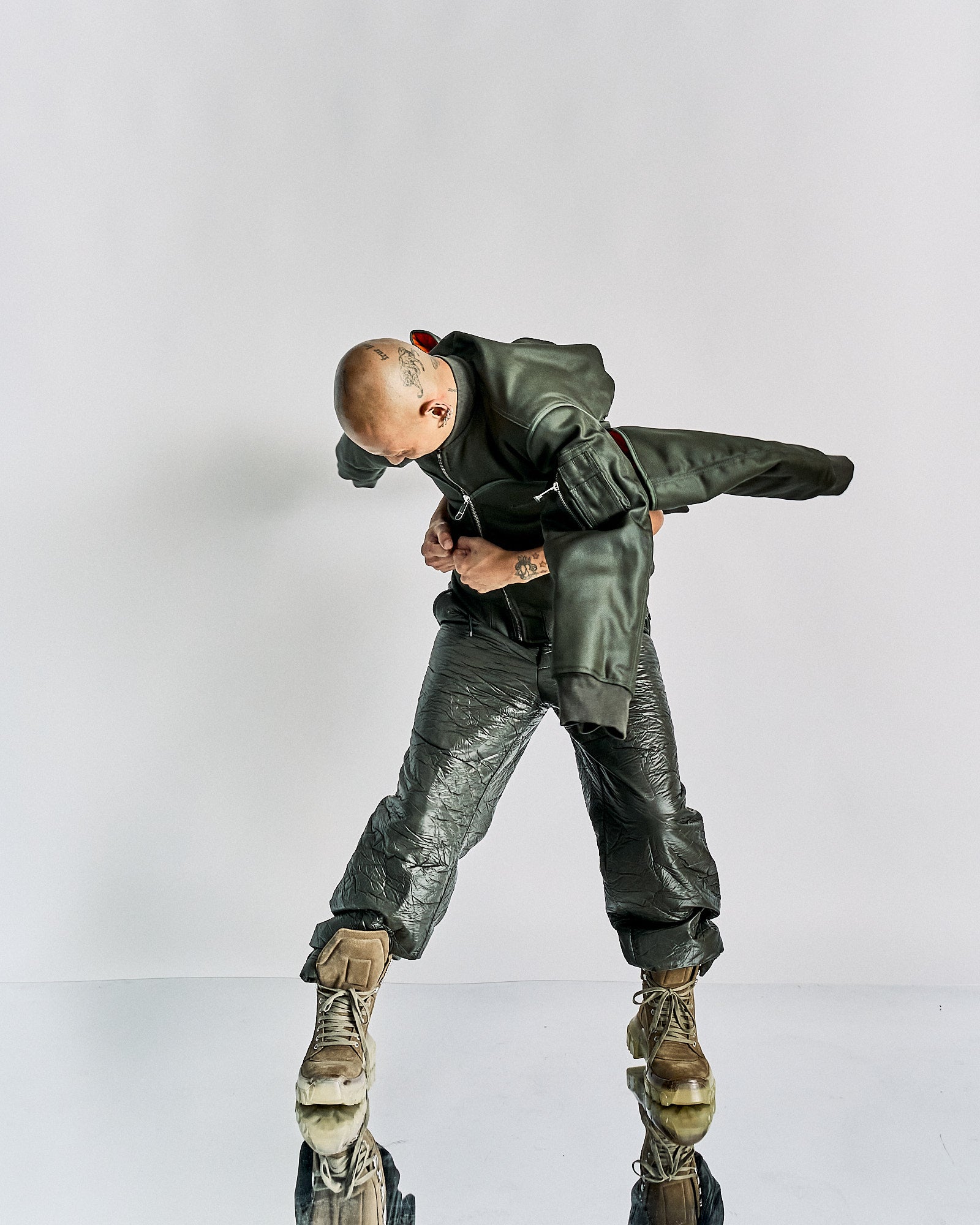A person in a futuristic outfit with a dark green Helmut Lang jacket and crinkled pants leans forward in a dynamic pose. They wear large brown boots. The background is plain white, emphasizing the avant-garde fashion style.