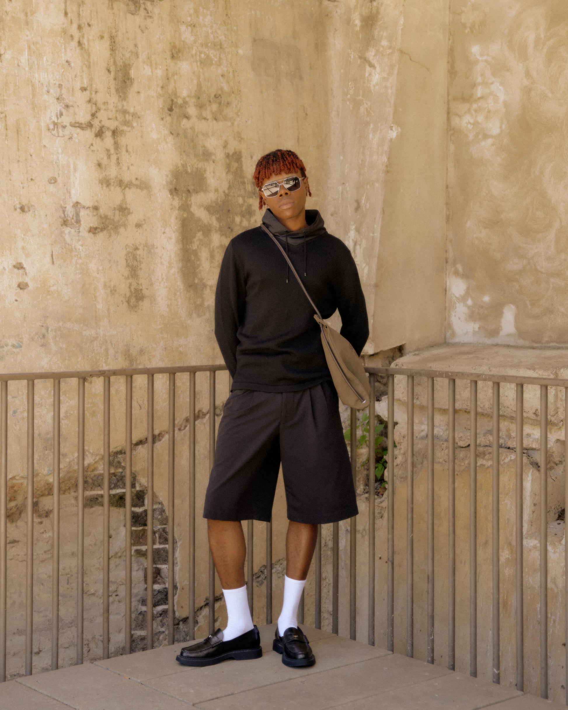 A person in a black Hermès hoodie and shorts leans against a textured wall. 