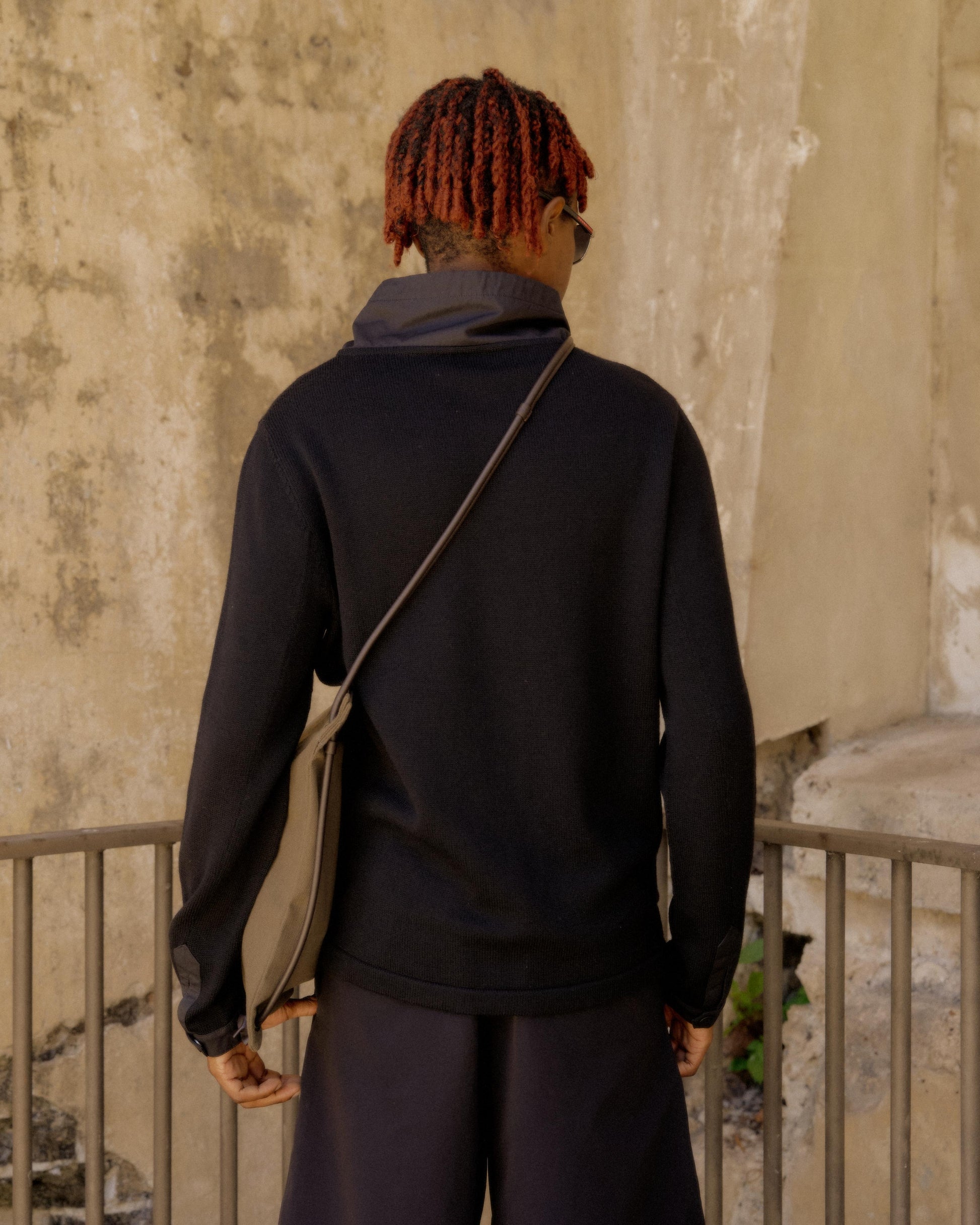 A person with red braided hair stands with their back to the camera, wearing a black Hermès sweater and carrying a grey shoulder bag. The background is a weathered beige wall.