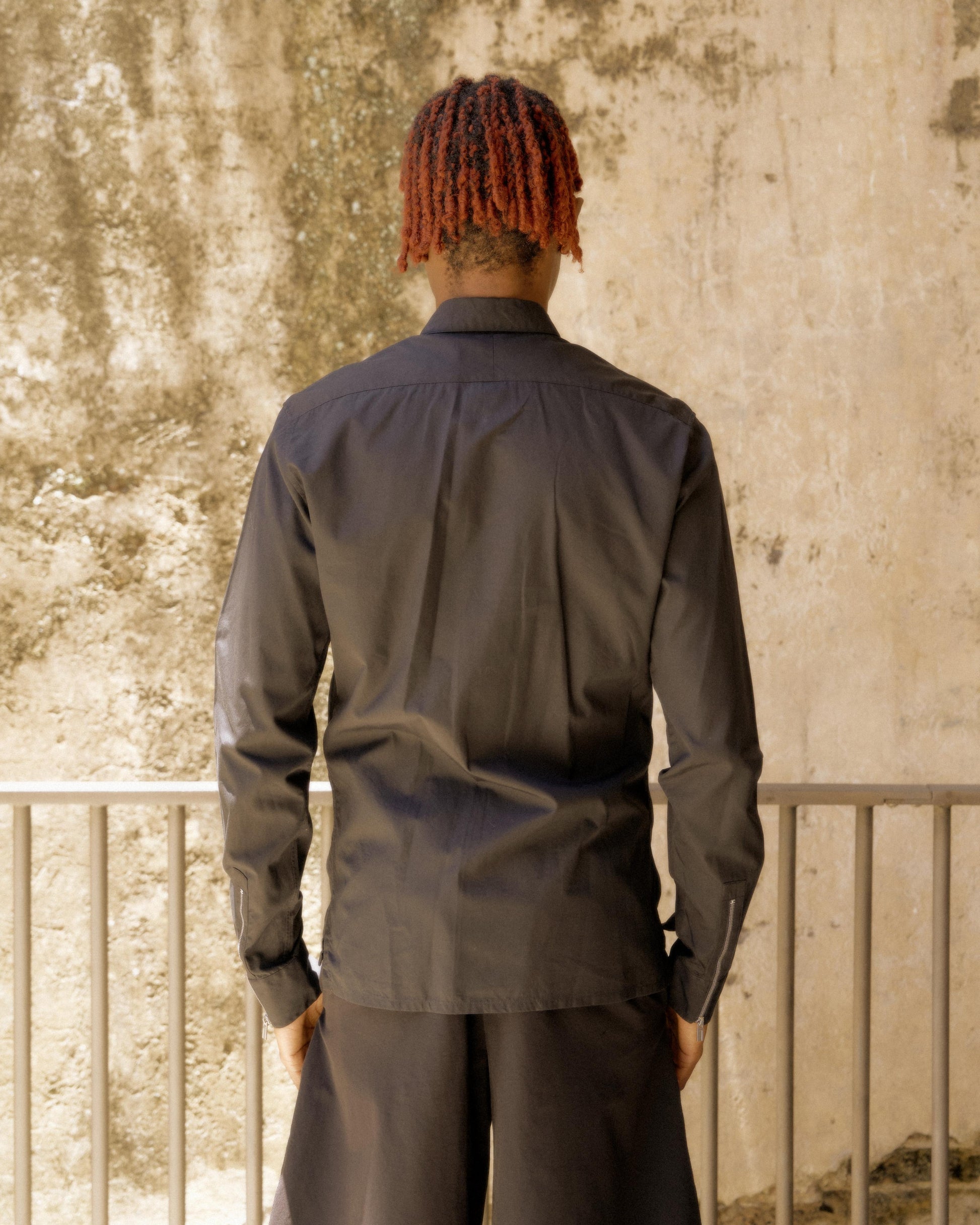 Person with red dreadlocks, seen from behind, wears a dark Hermès shirt and stands by a railing. The background is a textured, light-coloured wall.
