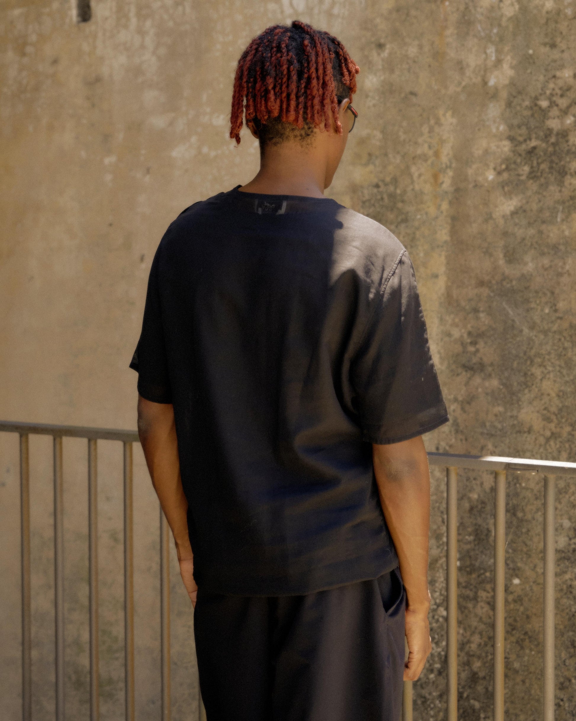A person with short red hair and sunglasses stands in profile, wearing a black Hermes outfit. 