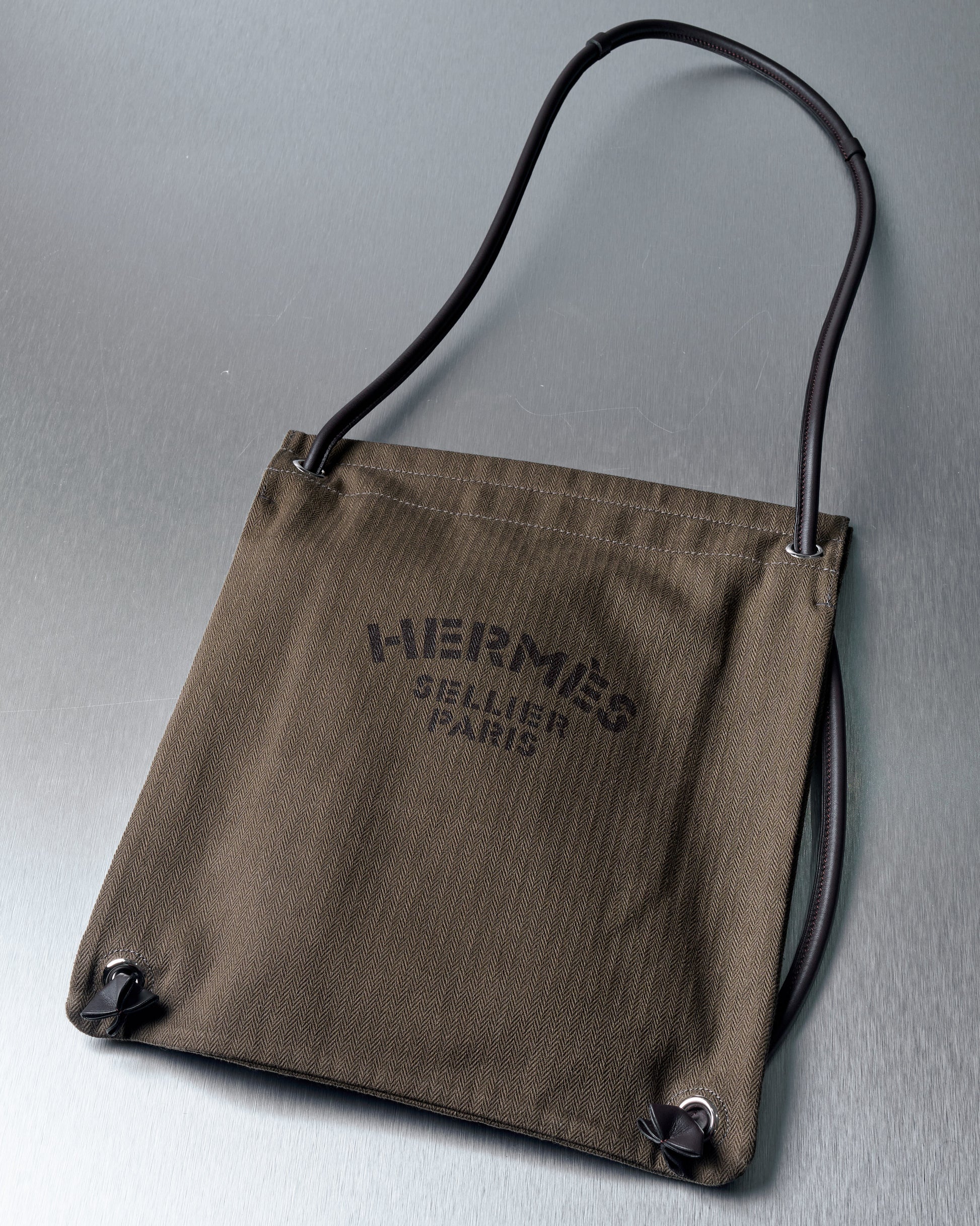 Olive green Hermes bag with black text "Hermes Sellier Paris" on the front, featuring black rope handles and a minimalist design on a metallic surface.