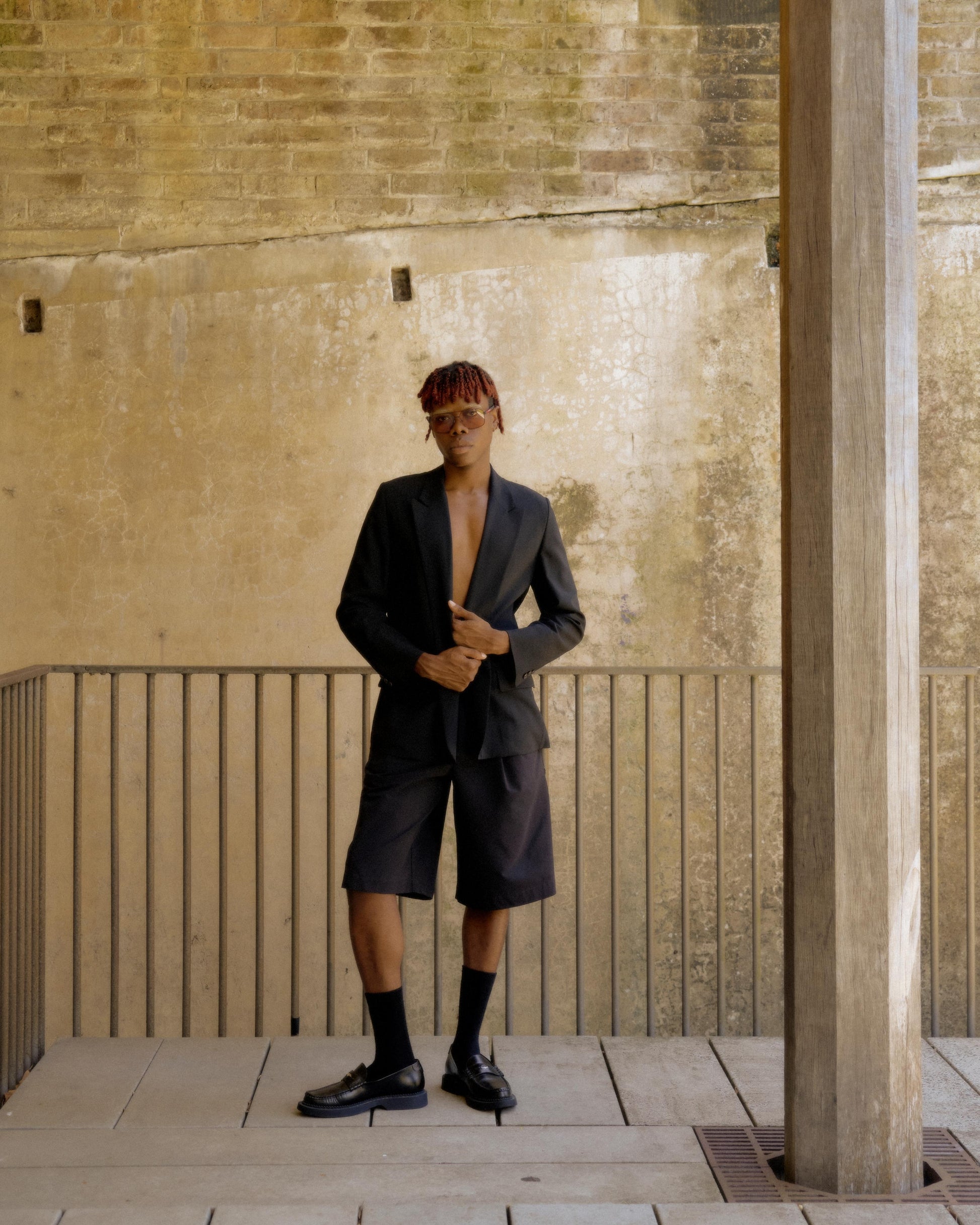 A person standing with one hand on a column, wearing a black long lapel blazer with a peak lapel, paired with black shorts and black shoes. The Hermès by Martin Margiela background is an indoor arched space with visible old bricks.