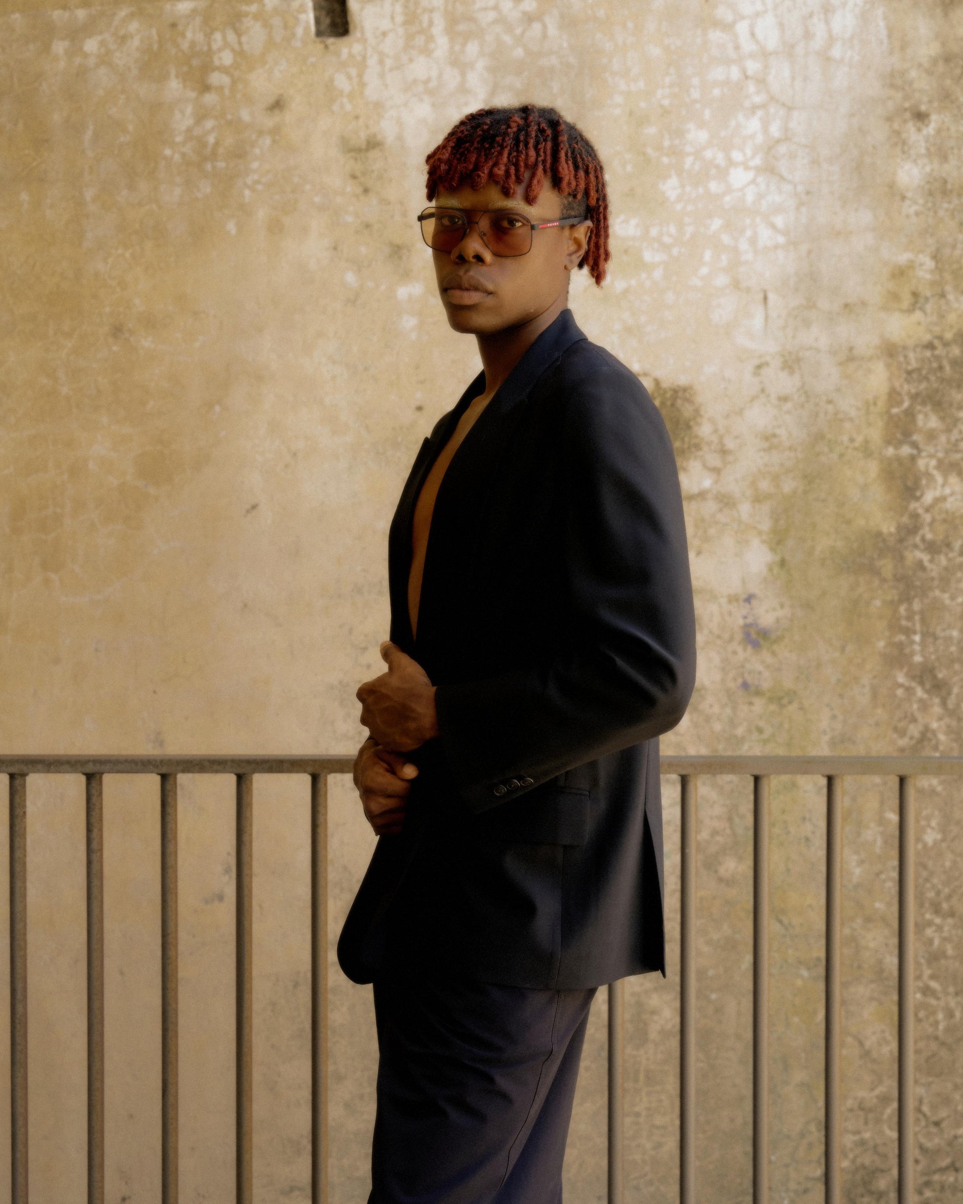 A person with red hair, wearing sunglasses and a dark Hermès suit, poses confidently against a textured beige wall