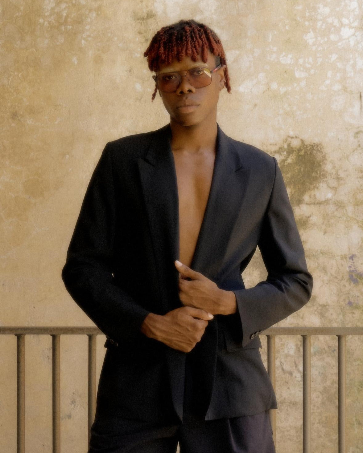 A person with red hair, wearing sunglasses and a dark Hermès suit, poses confidently against a textured beige wall, conveying a stylish, modern vibe.