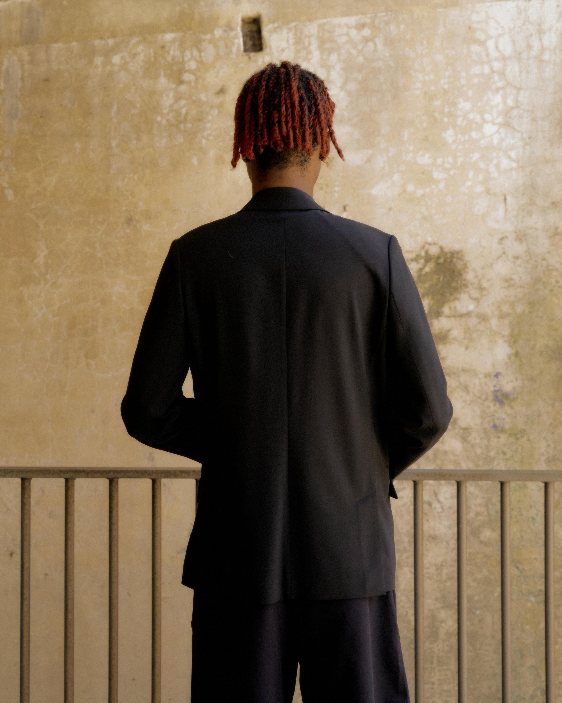 A person with red dreadlocks stands with their back turned, wearing a dark Hermès suit. They face an aged, textured wall, conveying a contemplative mood.
