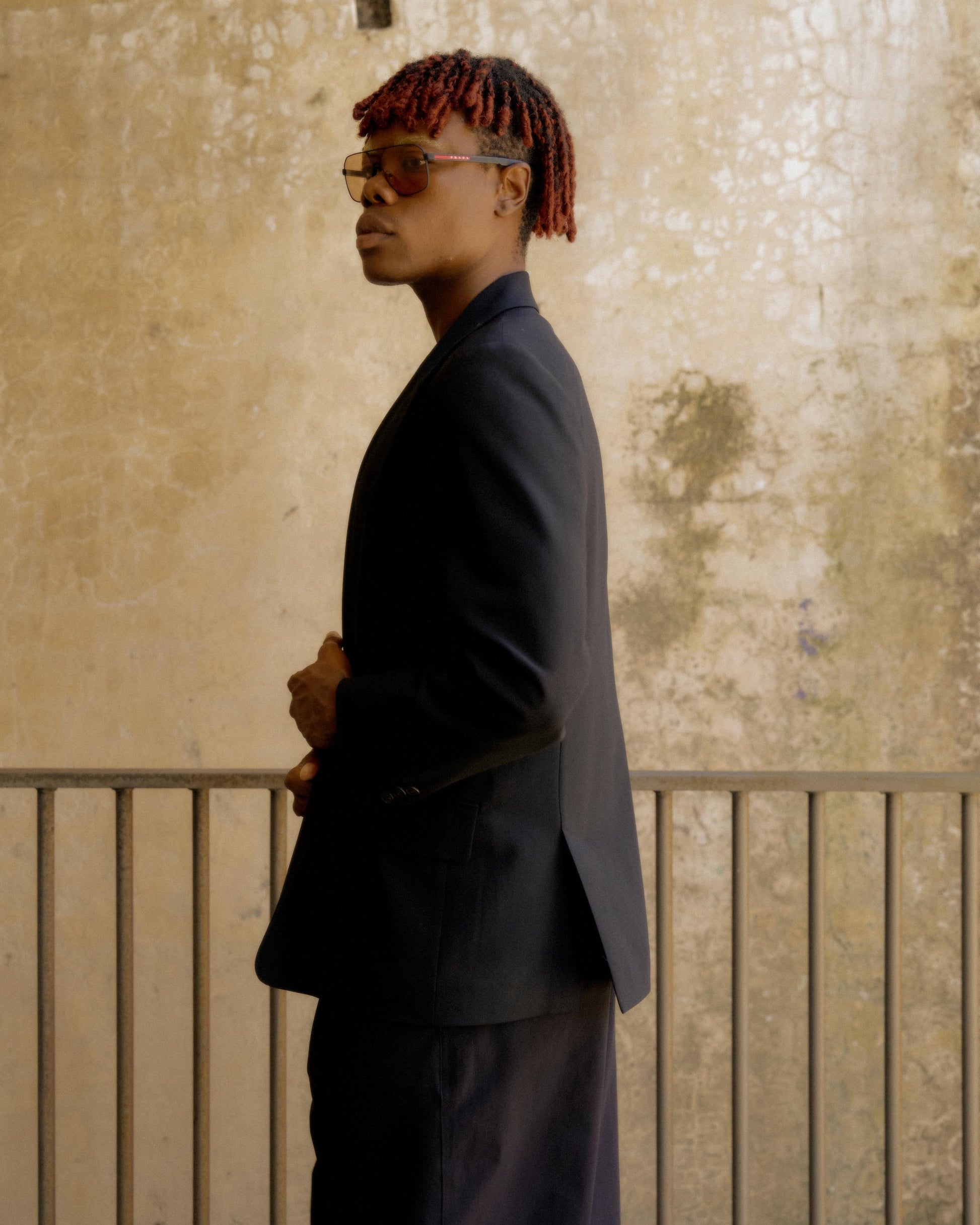 A person with short red locs, wearing glasses and a dark Hermès suit, stands sideways against a textured beige wall. 