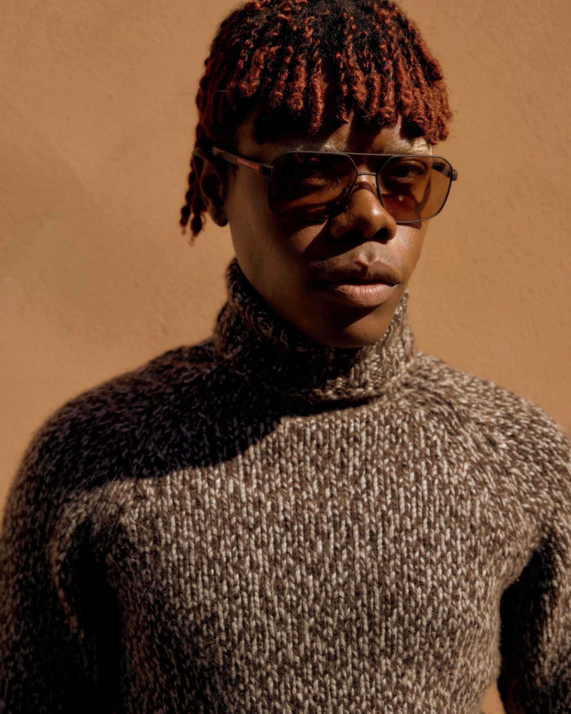 Person wearing sunglasses and a brown Hermes knit turtleneck stands against a warm tan wall. The image conveys a calm, stylish, autumn vibe.