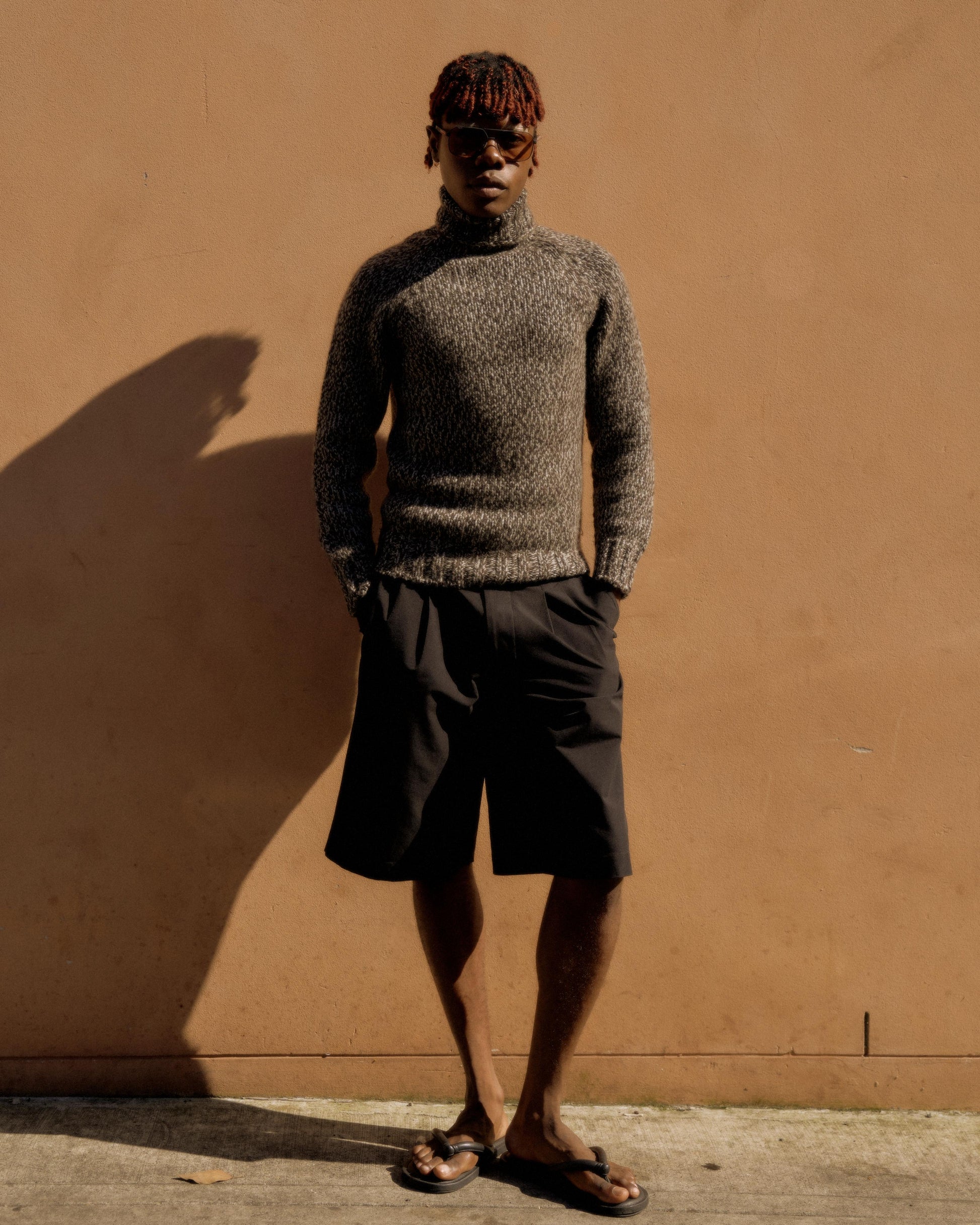 A person stands confidently against a tan wall, wearing a textured gray Hermes sweater, black shorts, and sunglasses. The mood is casual and relaxed, with warm lighting.