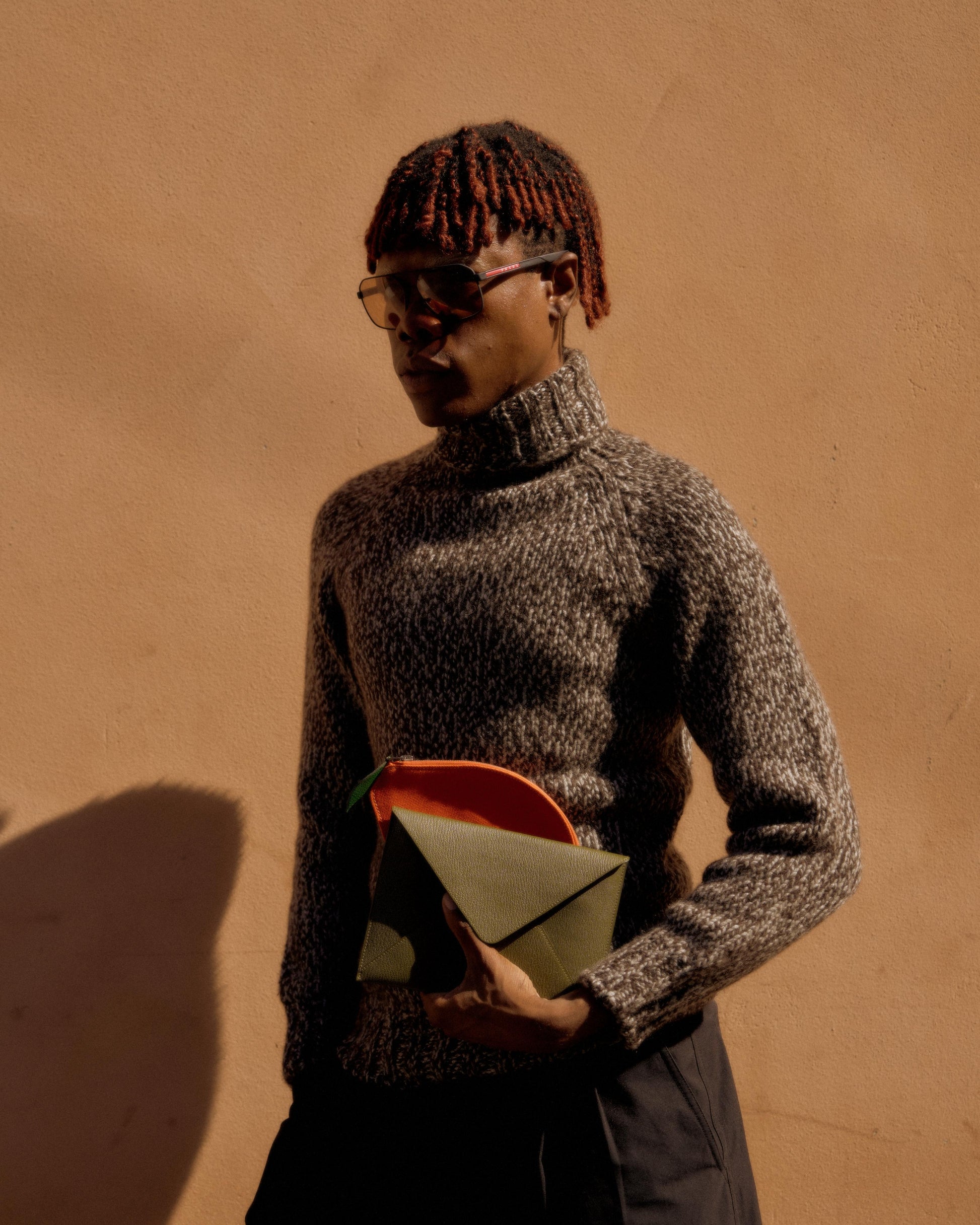 A person in a gray Hermes turtleneck sweater and sunglasses holds a multicolored clutch. They stand against a warm, peach-colored wall, casting a soft shadow.