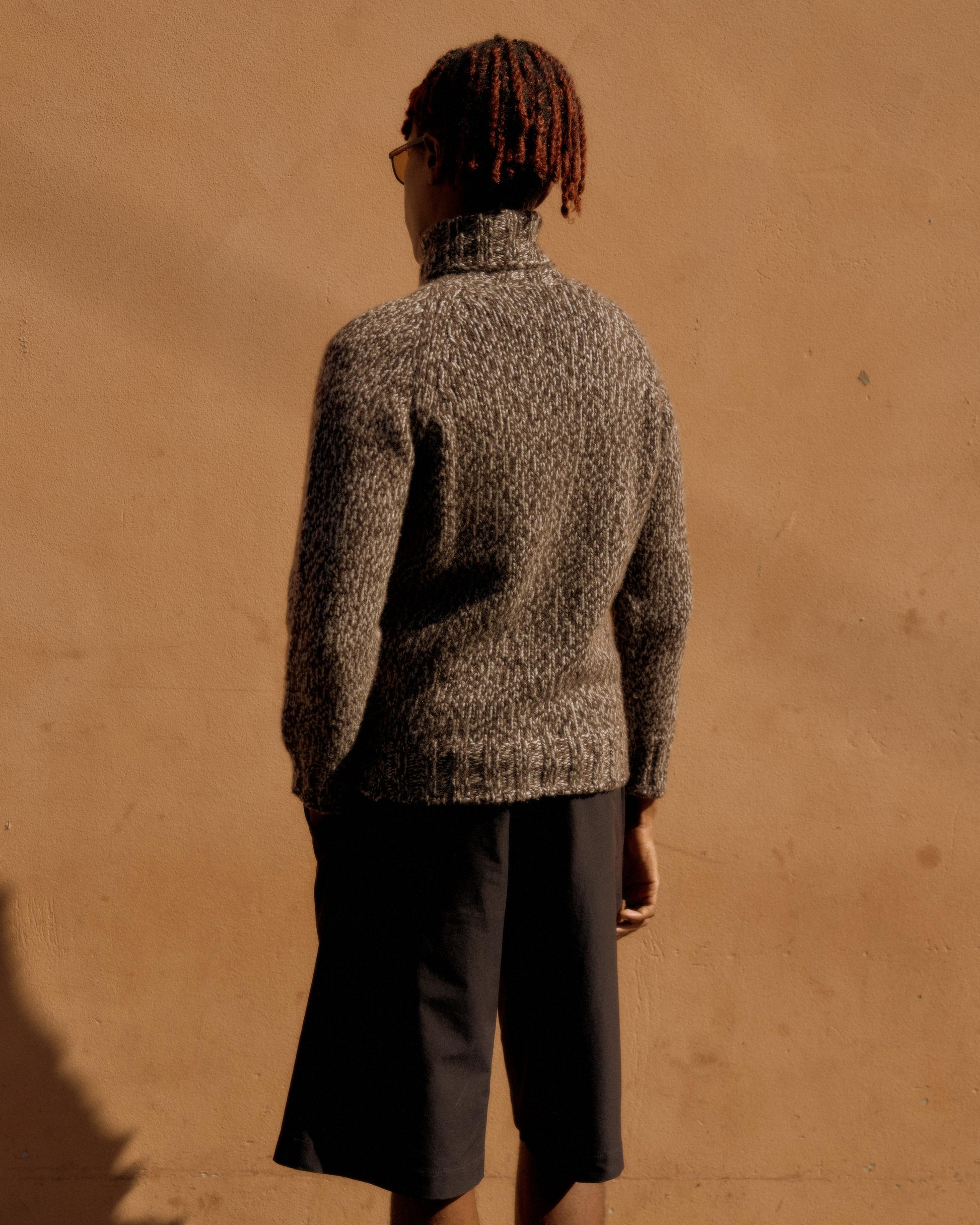 Person with red hair in cornrows and glasses, wearing a textured gray Hermes turtleneck sweater and black shorts, stands against a warm beige wall.