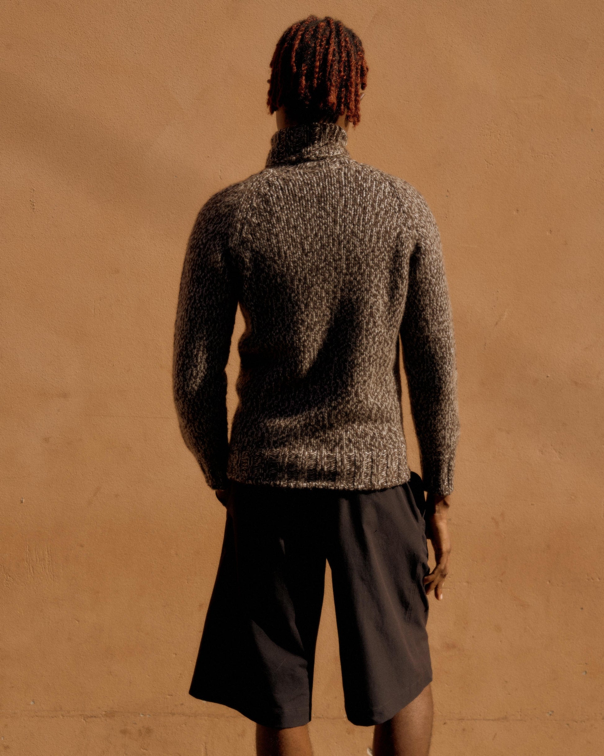 Person with red hair in cornrows and glasses, wearing a textured gray Hermes turtleneck sweater and black shorts, stands against a warm beige wall.