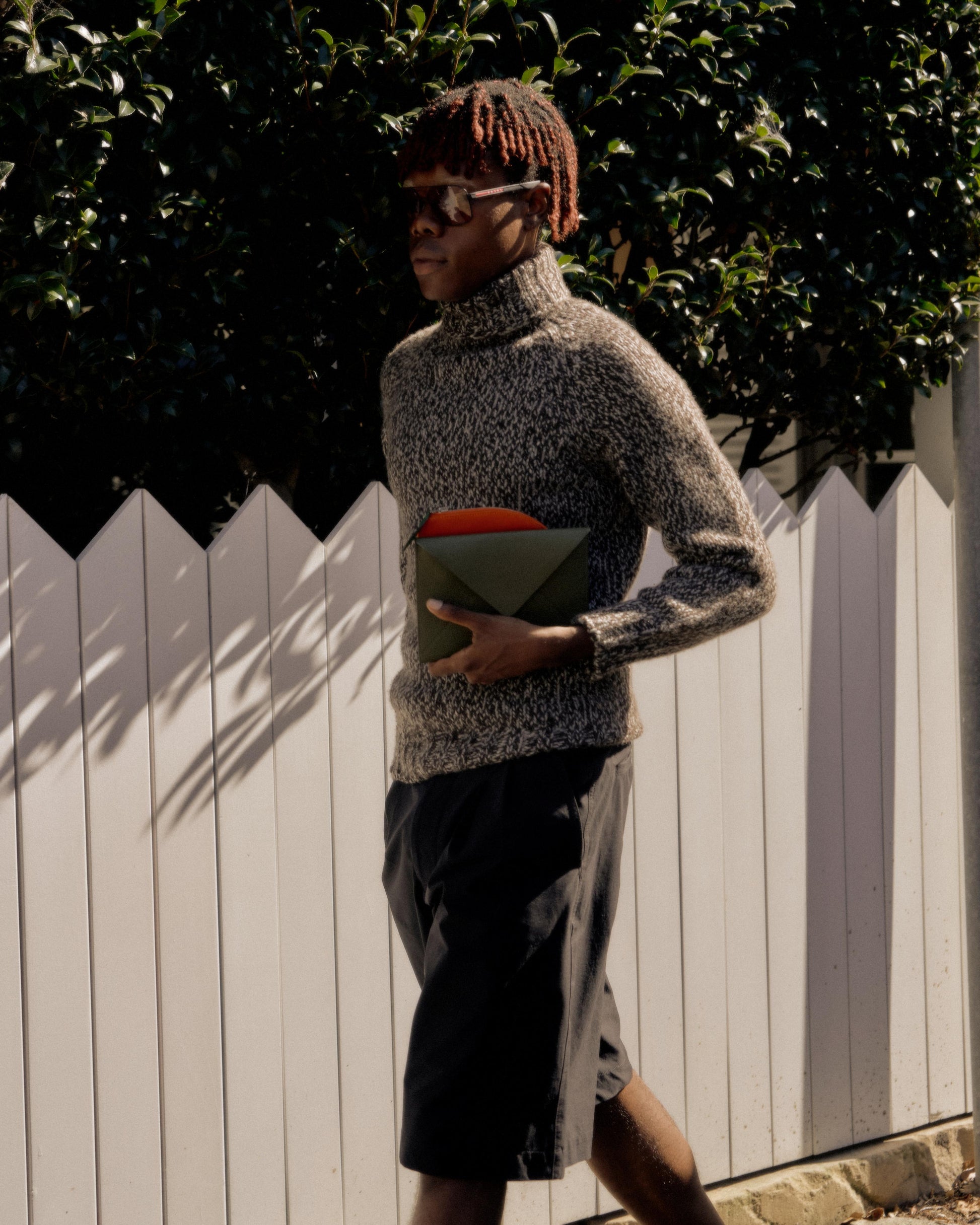 A person wearing a chunky marled Hermes cashmere turtleneck knit jumper with elongated roll over neck, raglan sleeves, rib knit cuffs and hem, carrying a red book, walking along a fence-lined path.