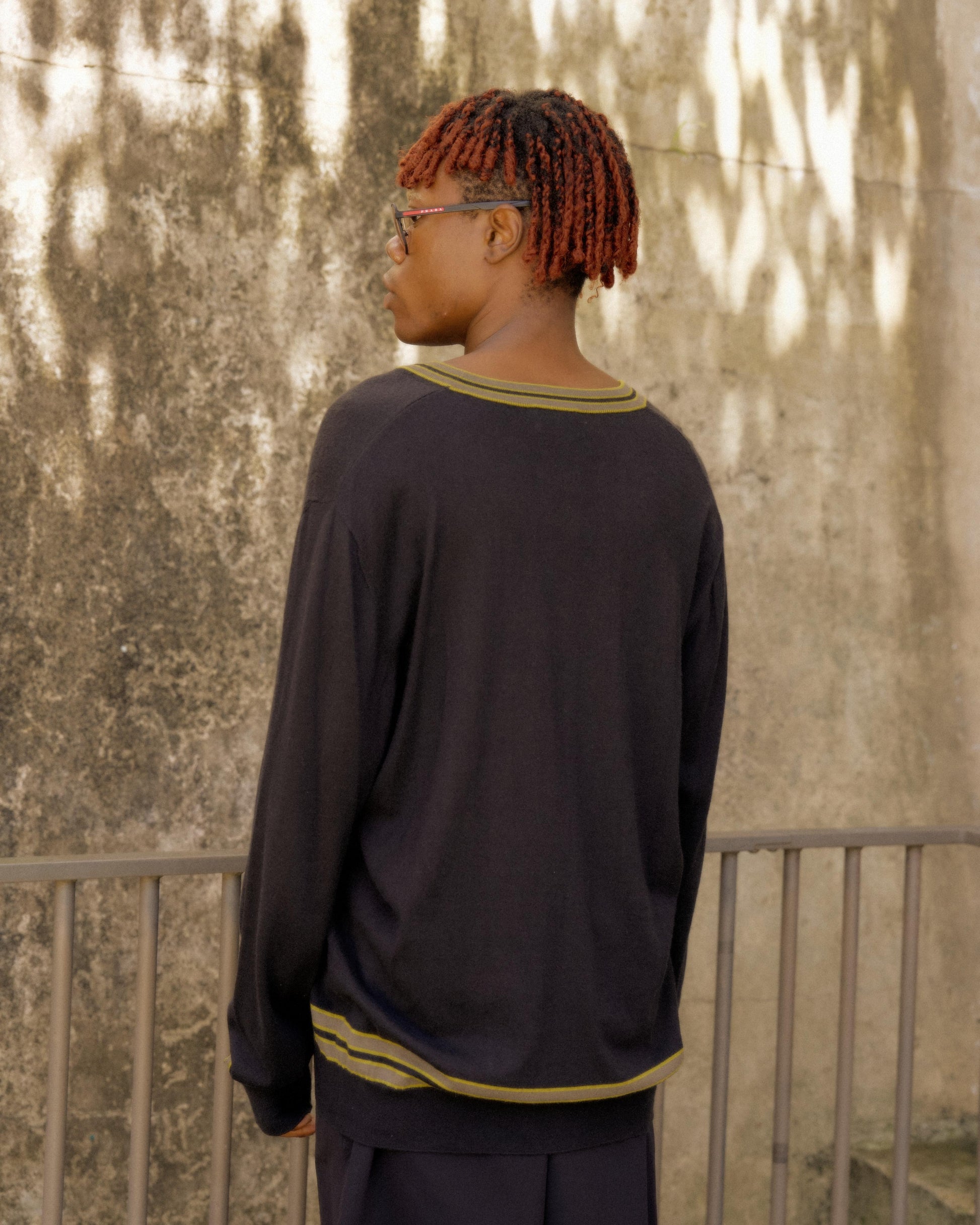 A person with short red dreadlocks stands with their back to the camera, wearing a black Hermes sweater featuring yellow stripes. A textured stone wall is in the background.