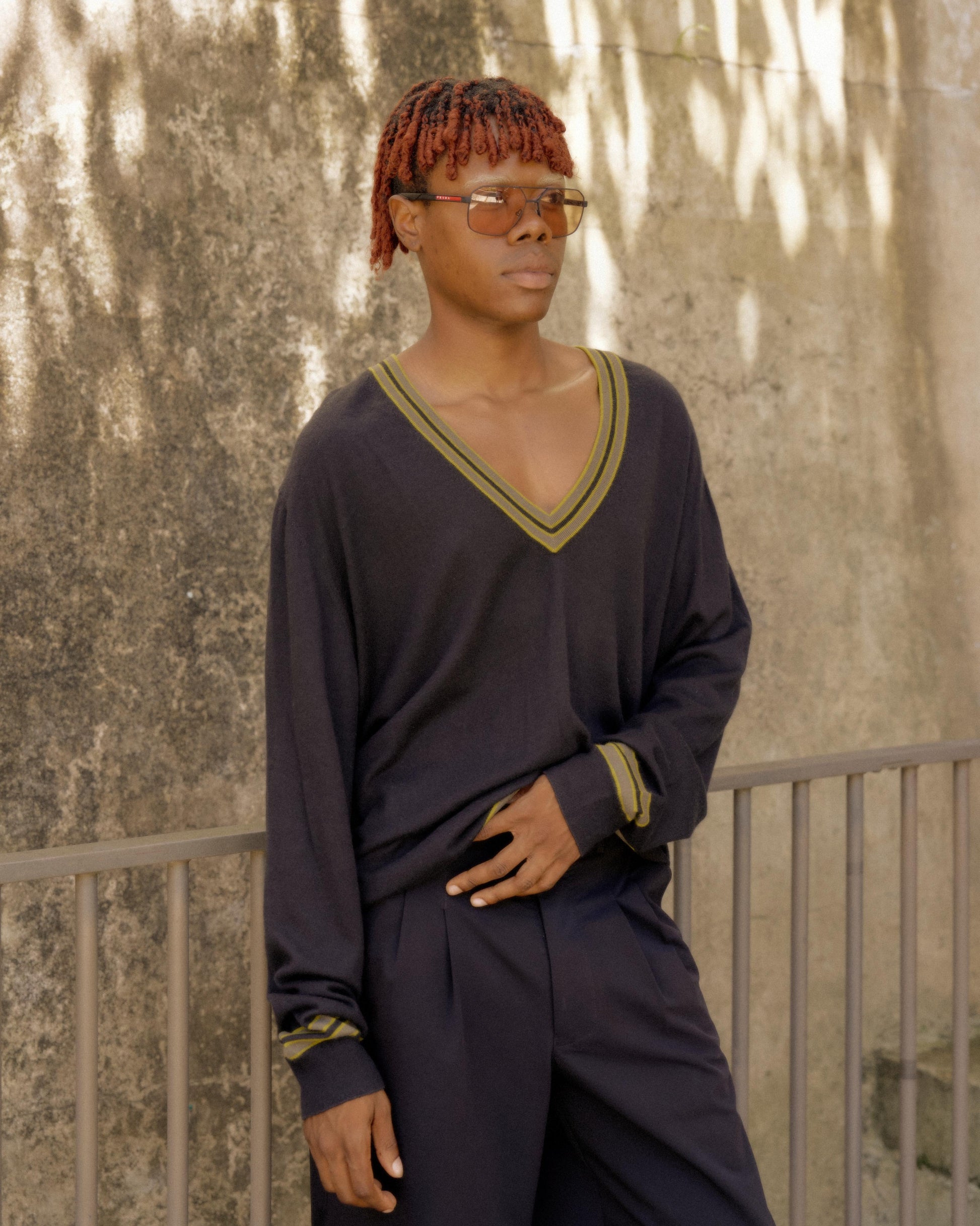 Young person with sunglasses and orange braids wears a dark Hermes V-neck sweater and matching pants, leaning against a rail, with a textured wall background.