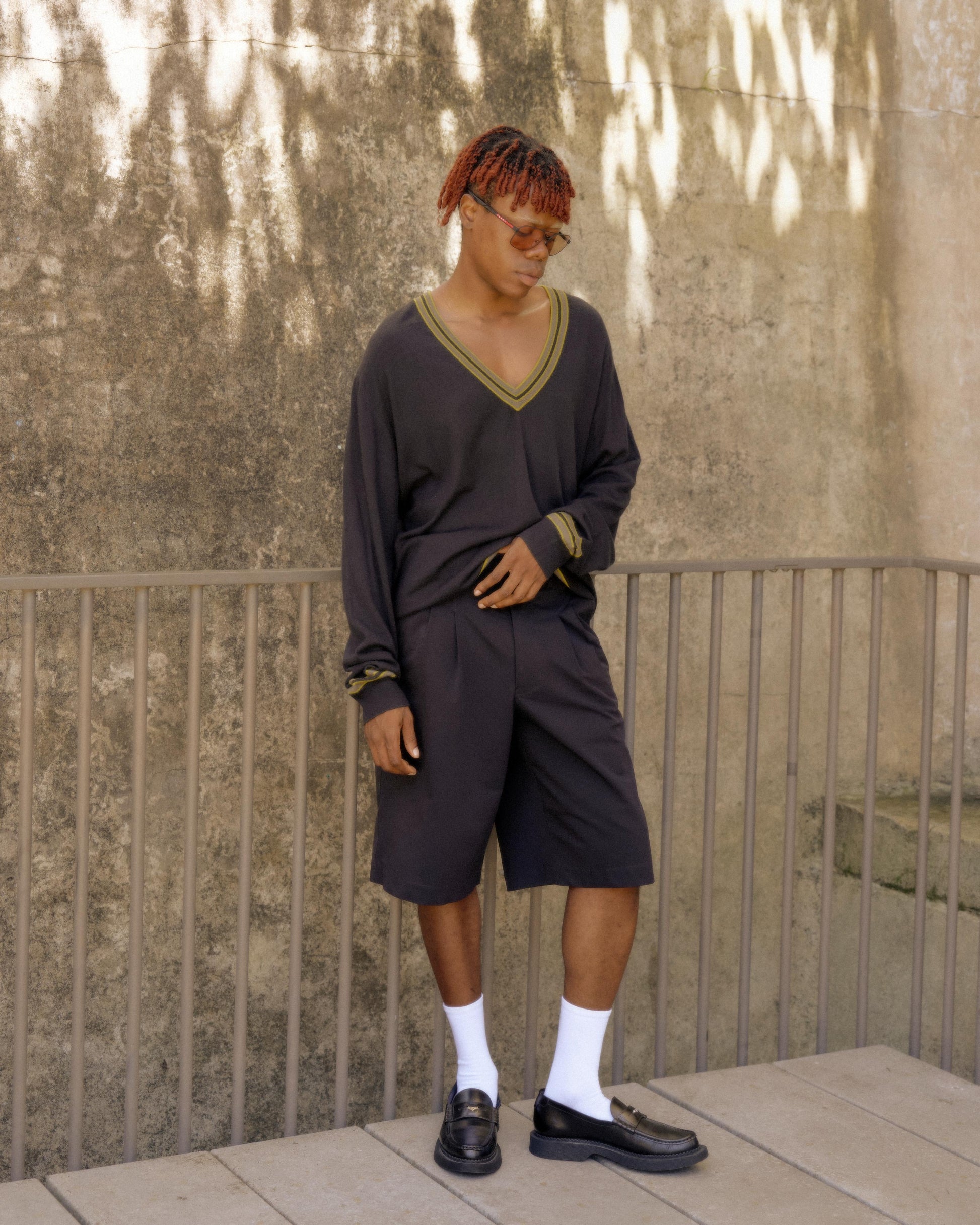 A male model wearing a V-neck Hermes knit cashmere sweater and shorts, standing against a textured background.