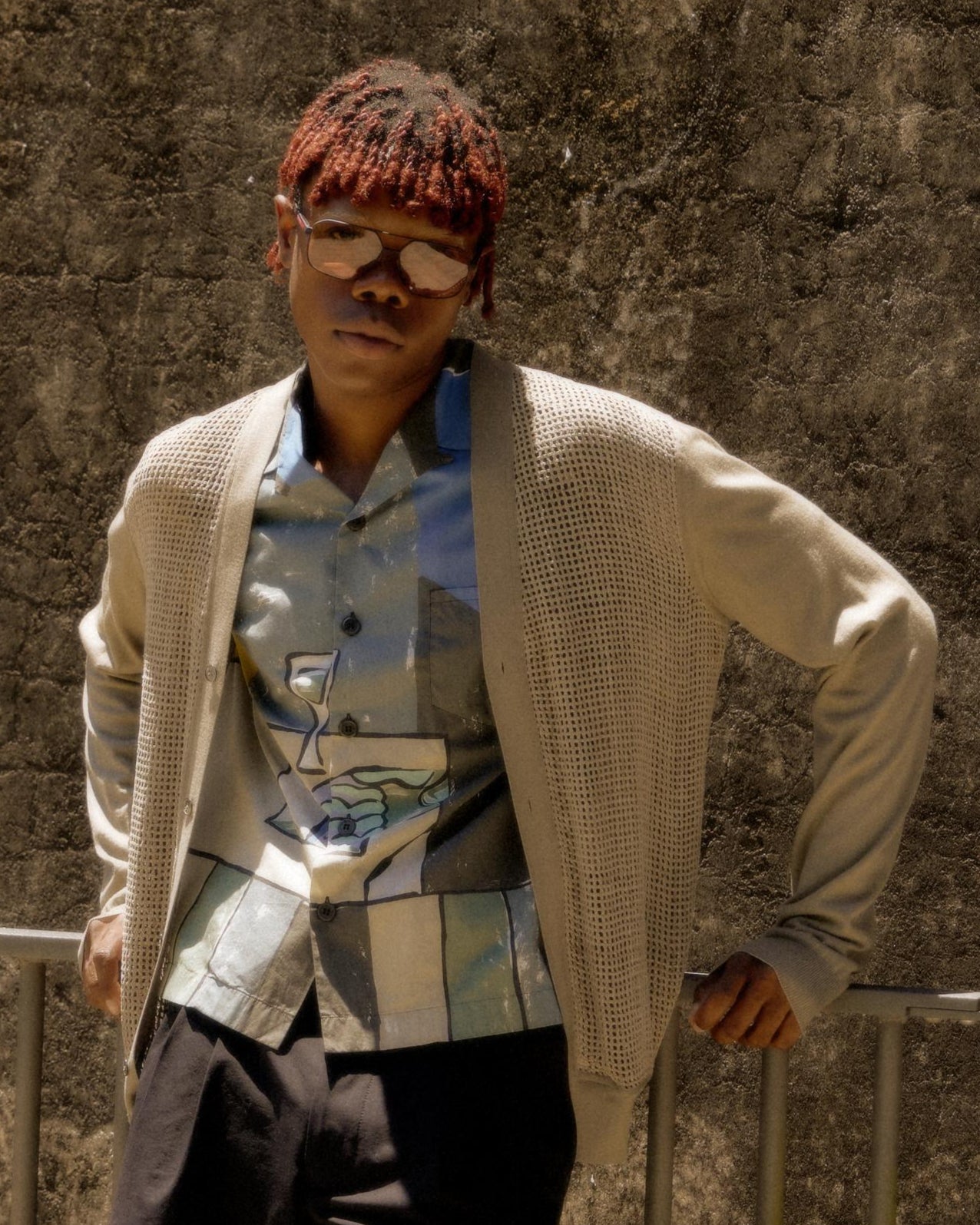 A person with red braids wears sunglasses, a light Hermès cardigan, and a patterned shirt while leaning against a concrete wall, exuding a relaxed, confident vibe.