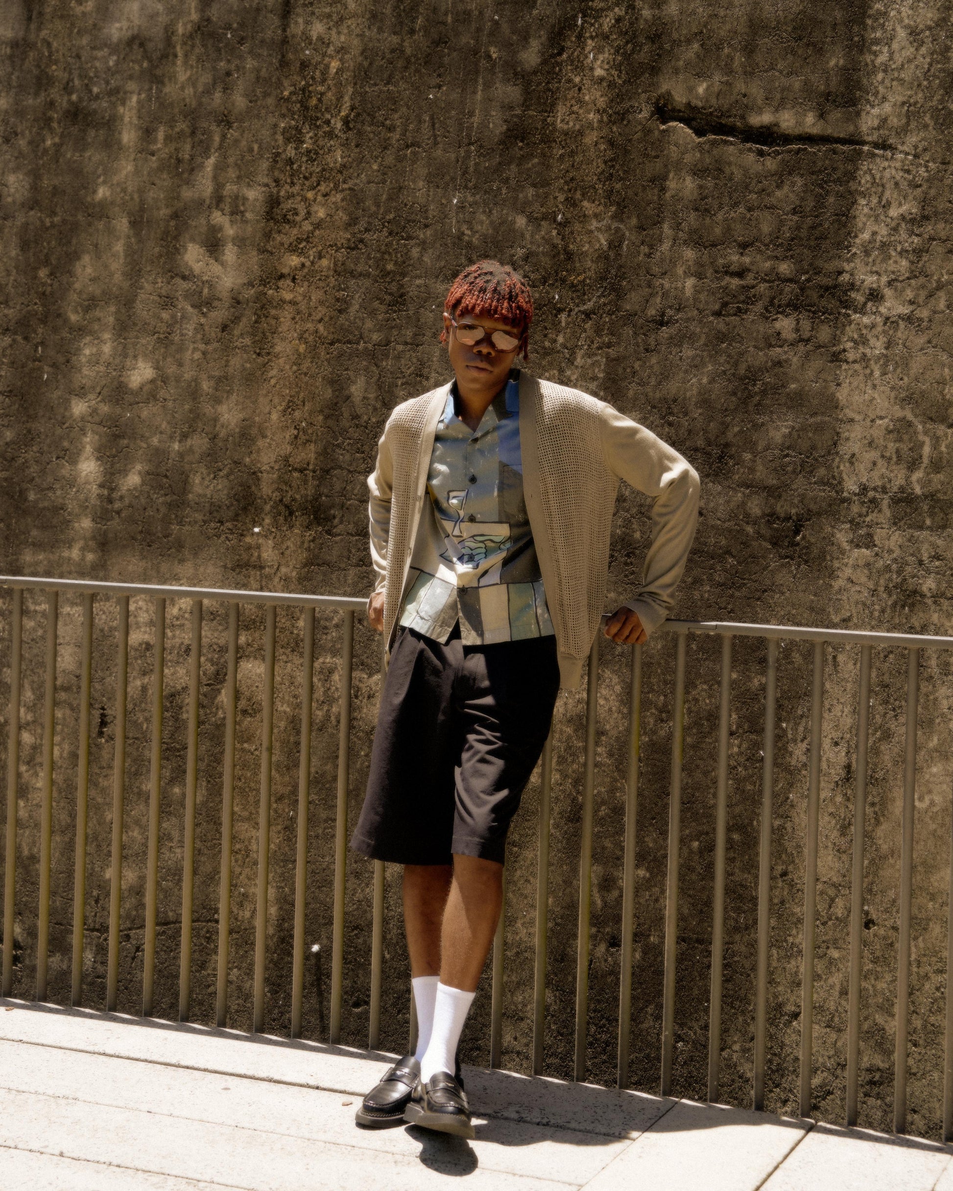 Man leans casually against a metal railing in sunny outdoor setting. Wears sunglasses, patterned shirt, beige Hermes cardigan, black shorts, white socks, and loafers. 
