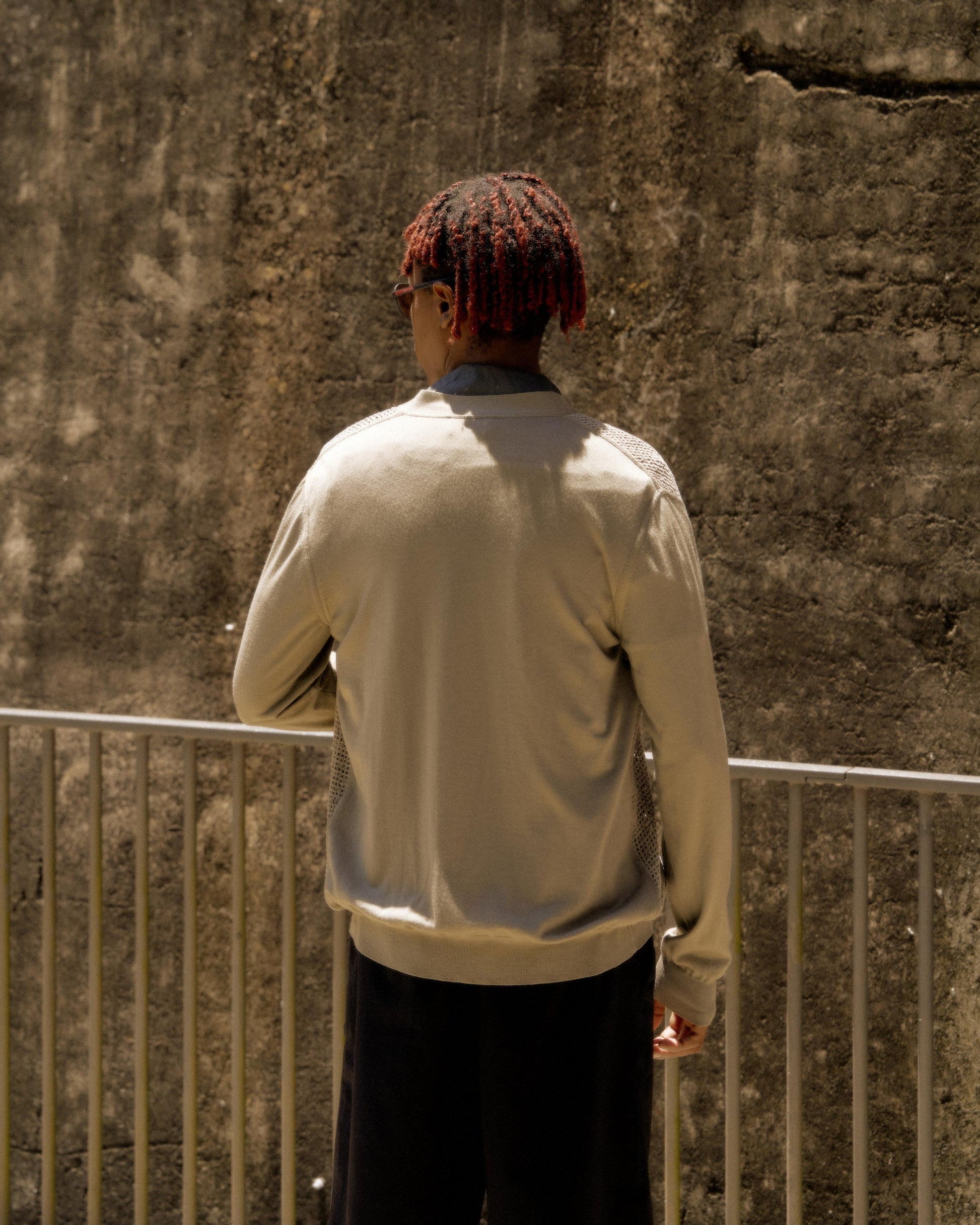 A person with braided red hair stands by a concrete wall, wearing a light Hermes sweater and dark pants. They face away, evoking a contemplative mood.