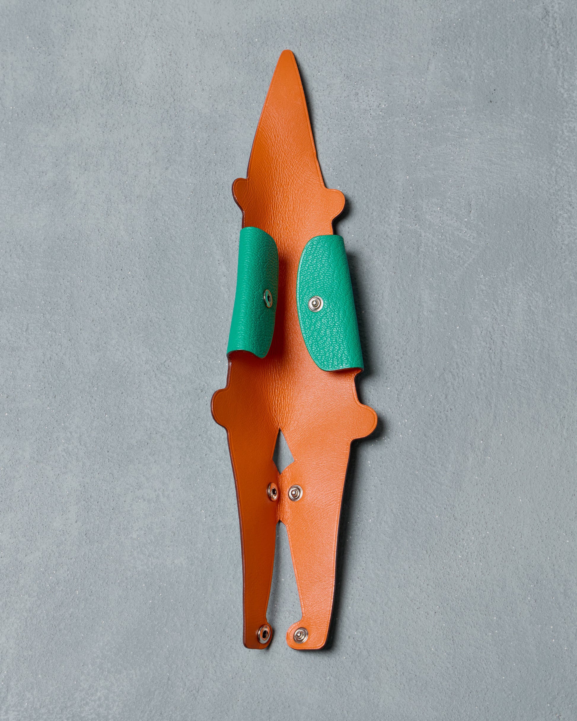 Green and orange crocodile-shaped art object from Hermes with silver rivets, set against a textured gray background. Sleek and playful design, exuding a whimsical tone.