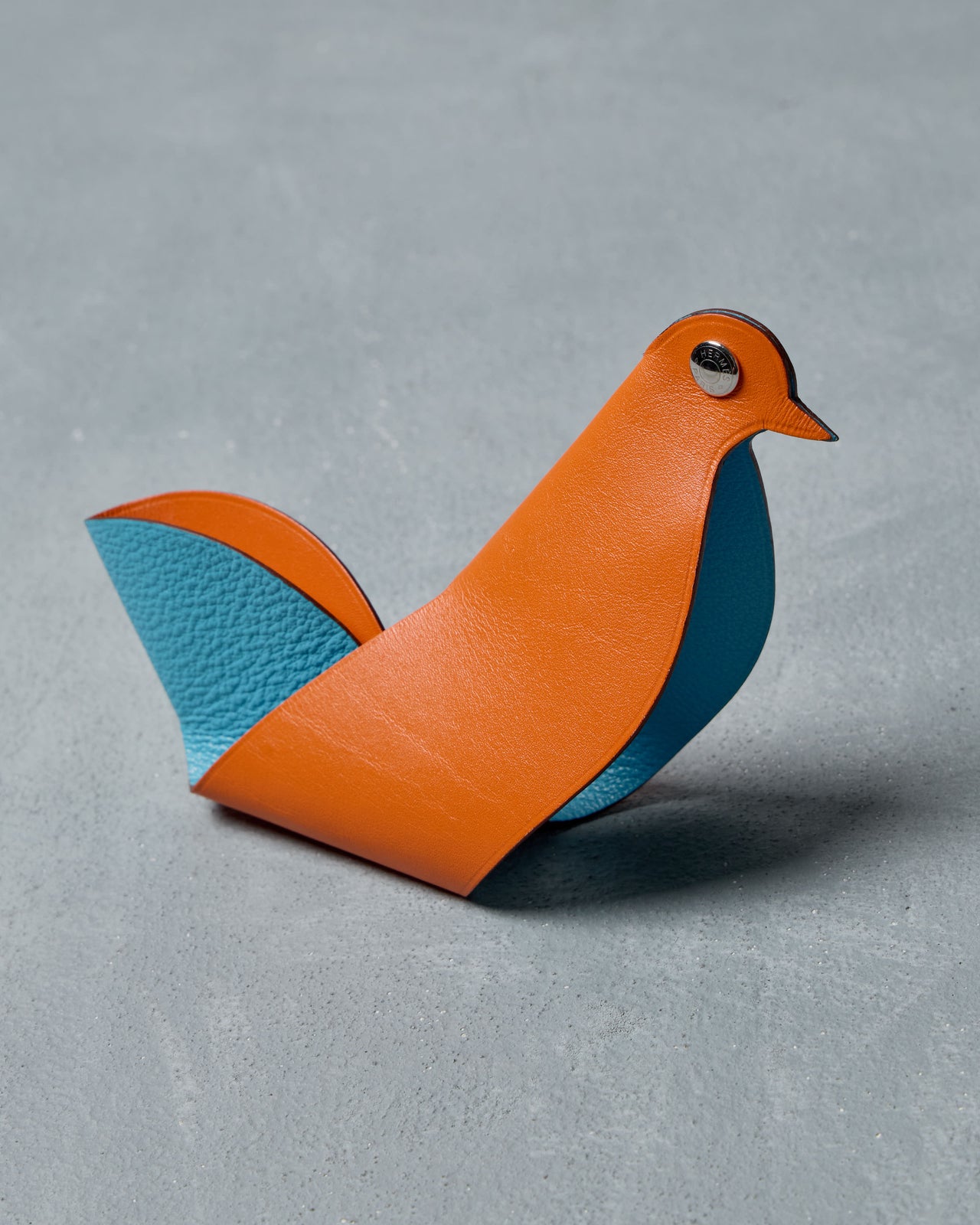 A minimalist, abstract bird sculpture from Hermès made from orange and blue leather, featuring a shiny metal eye, on a textured gray surface. Modern and playful design.