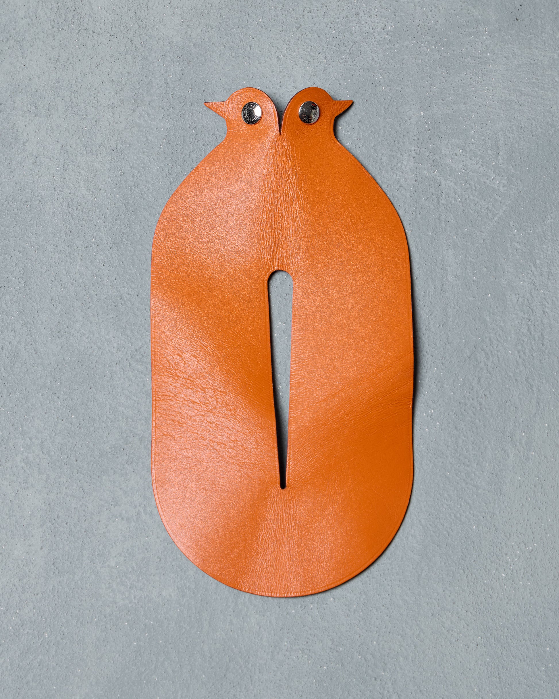 Orange leather Hermès piece resembling two stylised birds facing each other, with metallic eyes, placed against a grey textured background.