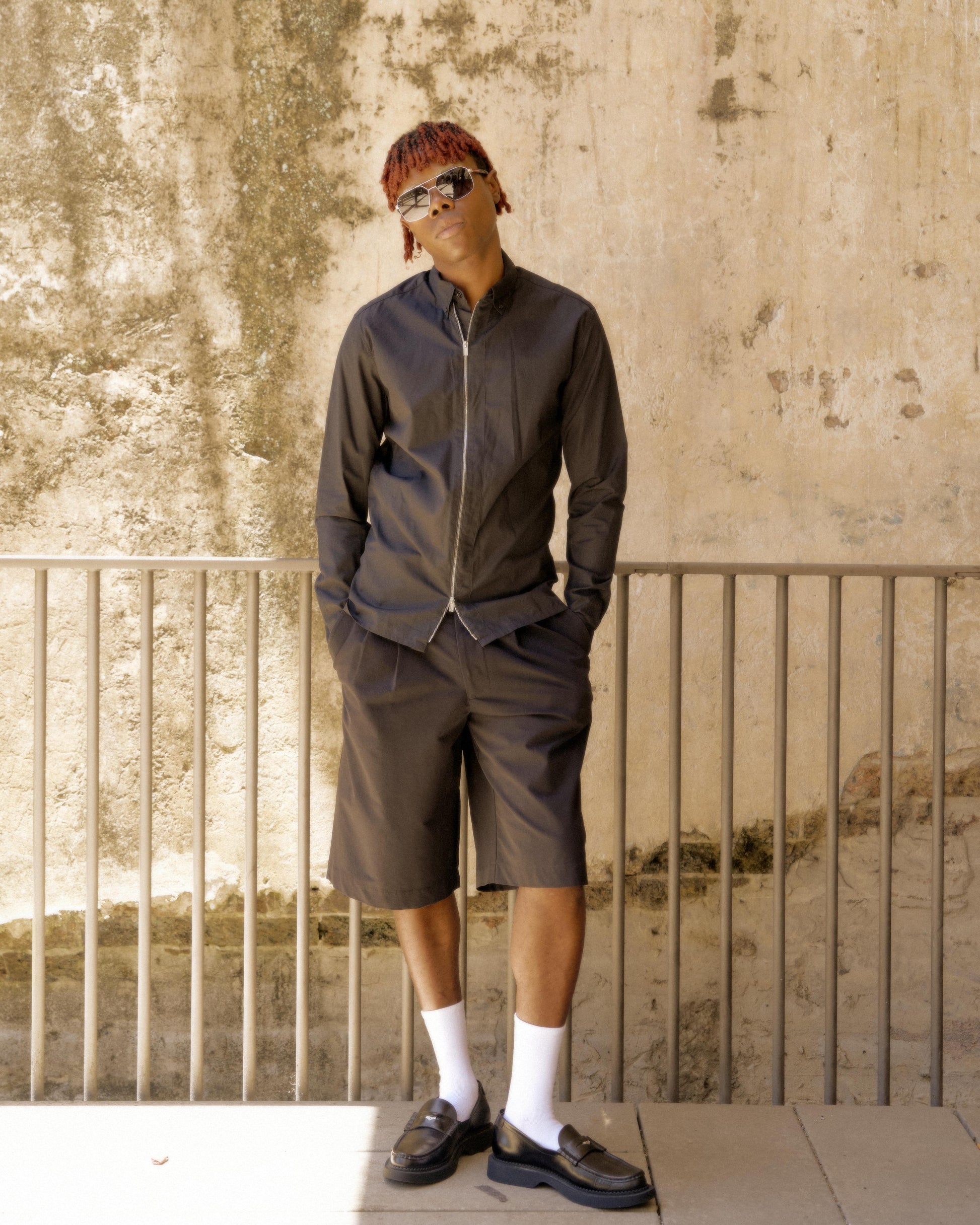 Stylishly dressed person leans casually against a textured wall, wearing sunglasses, dark Hermès shirt and shorts, with white socks and loafers