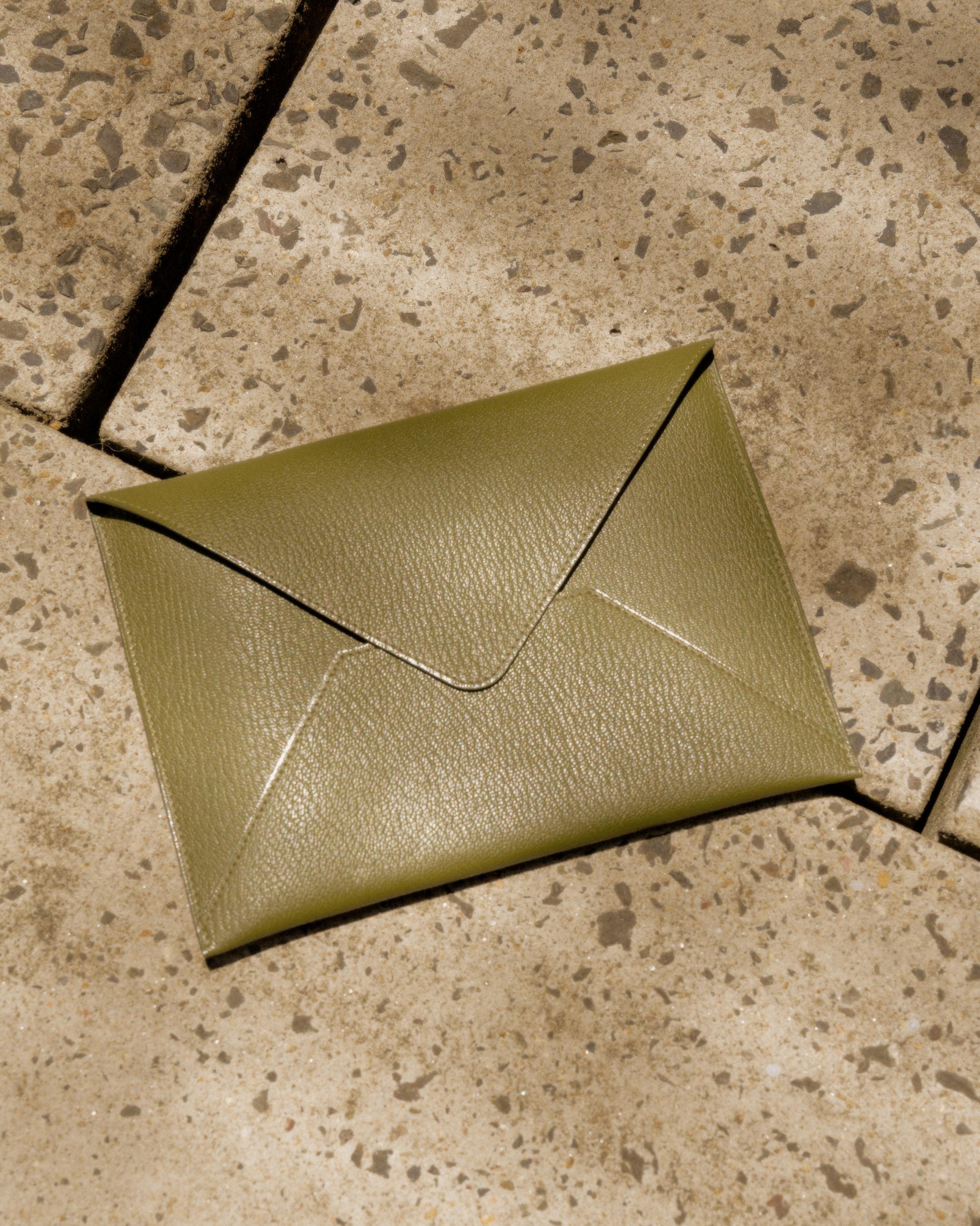 A green leather Hermès envelope clutch lies on a textured stone surface, casting a soft shadow.