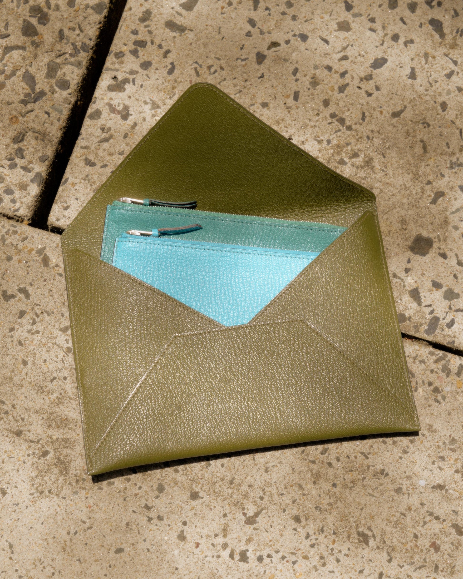 A green leather Hermès envelope contains a vibrant turquoise wallet with silver zippers, resting on a textured stone surface in sunlight.