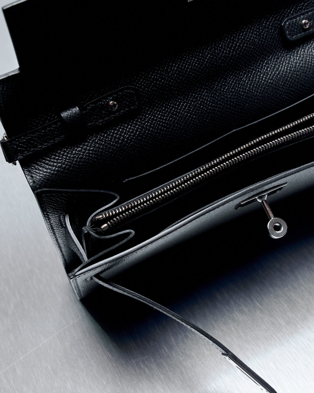 Sleek black leather Hermes clutch with a long strap on a brushed metal surface. Features a silver clasp.