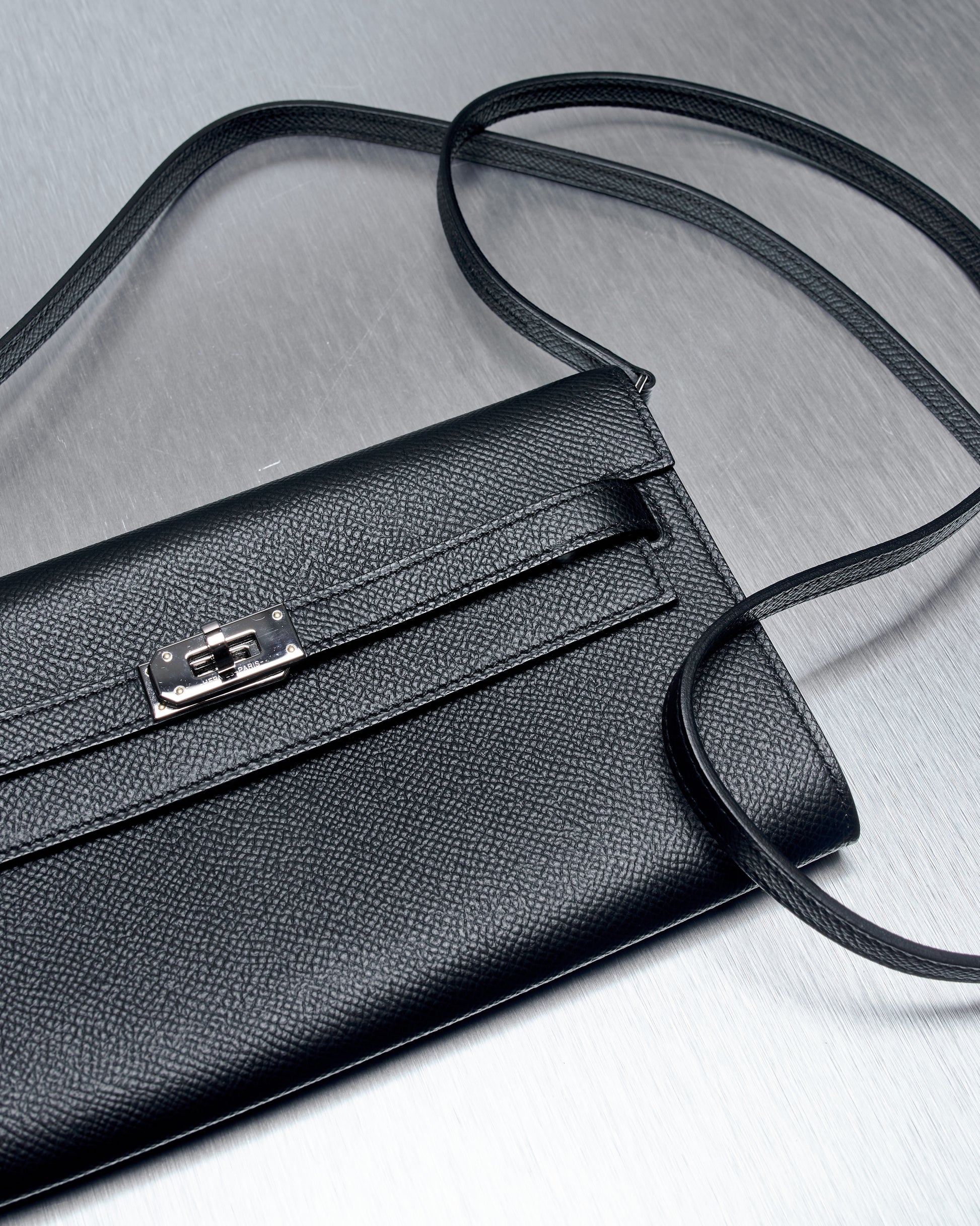 A sleek black leather Hermès purse with a textured surface and elegant silver clasp, laid on a brushed metal surface