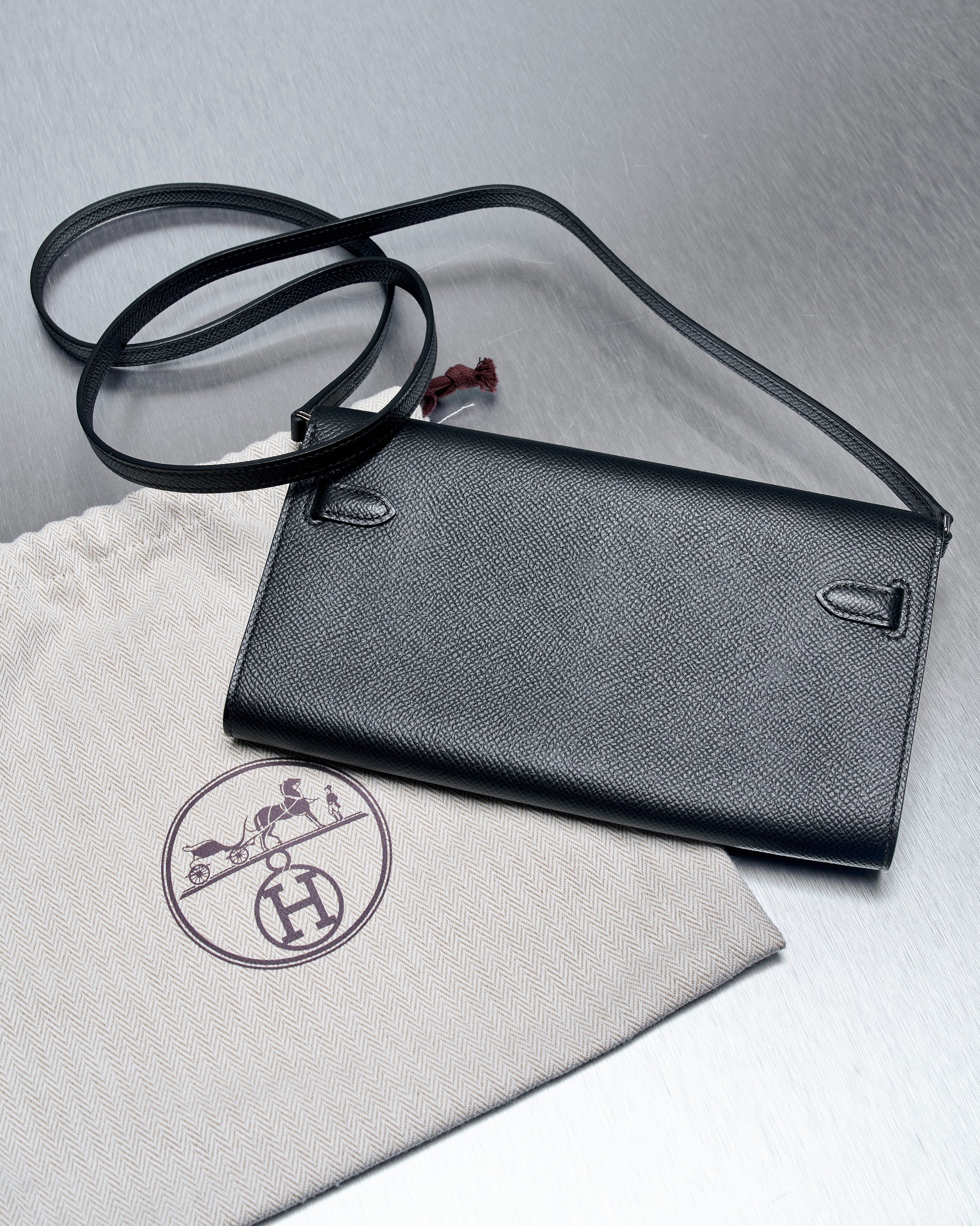 Black leather Hermès clutch with a long strap on a metallic surface next to a beige fabric pouch featuring a logo. 