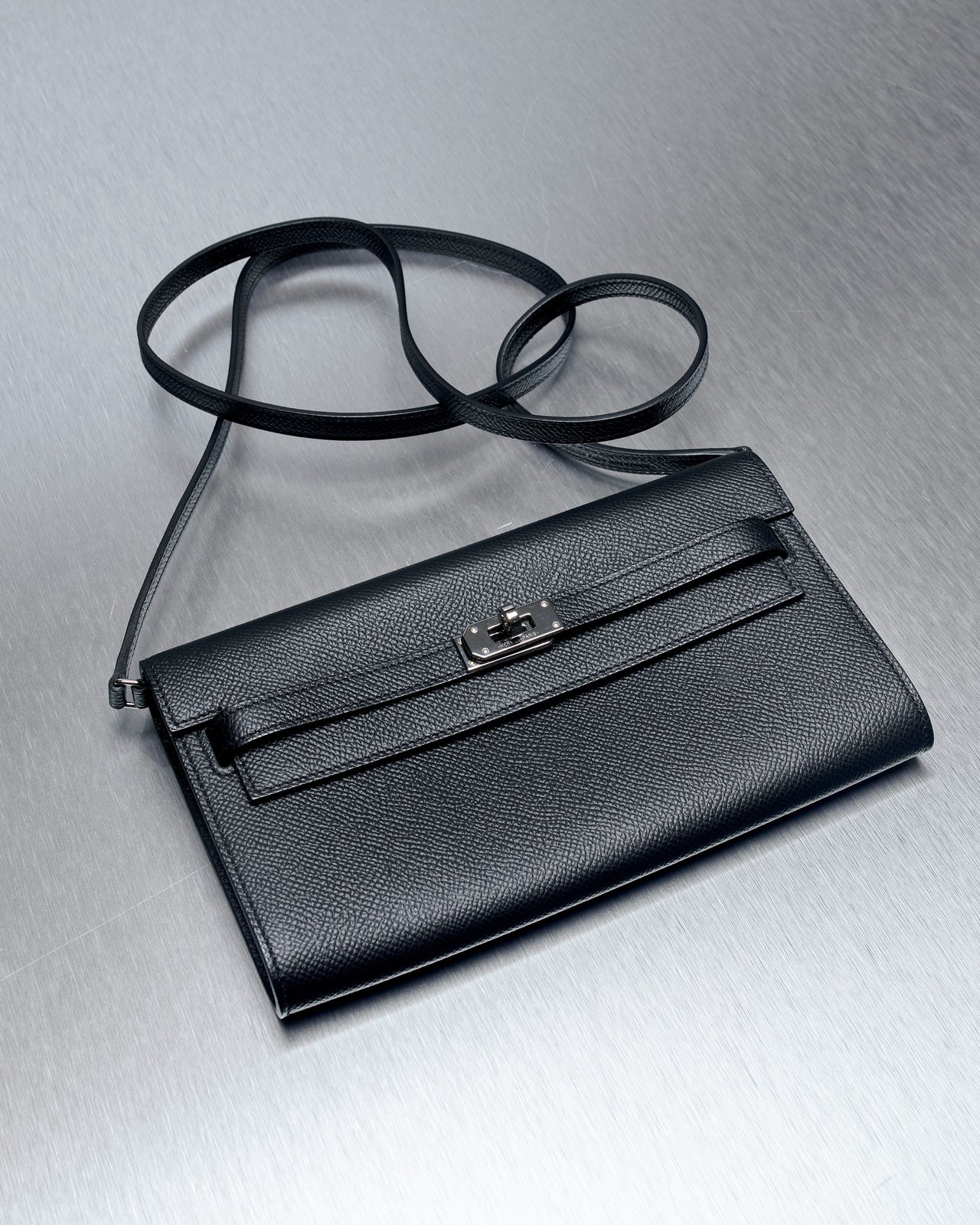 Sleek black leather Hermes clutch with a long strap on a brushed metal surface. Features a silver clasp.