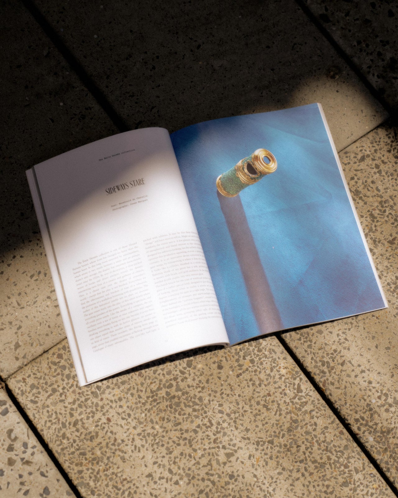 A magazine from Hermès  titled "Le Monde" lies on a sunlit, textured pavement. The cover features abstract art in bright colors. The scene feels casual and serene.