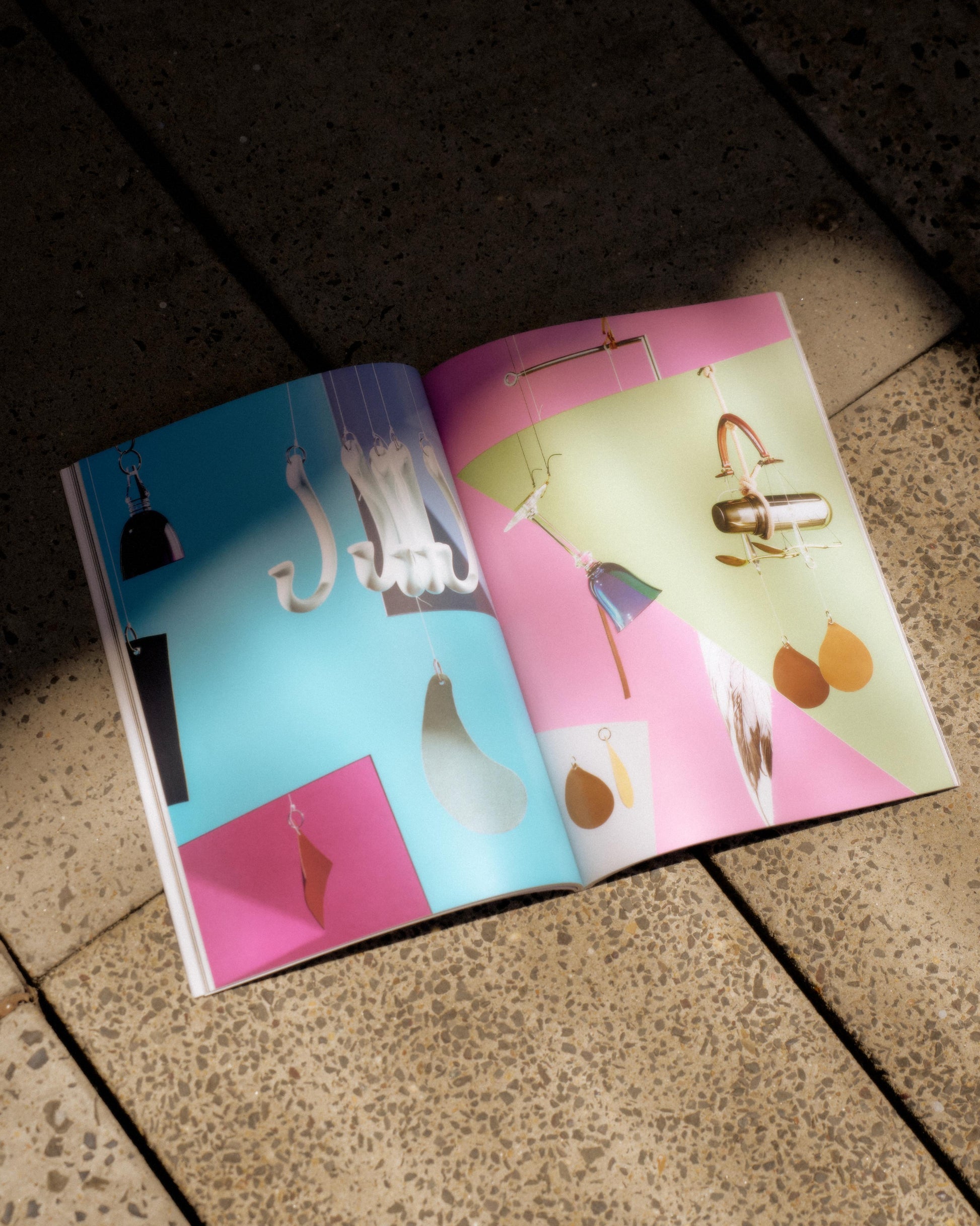 An open Hermes magazine on a stone floor displays colourful geometric pages with hanging jewellry, including earrings and hooks, on blue and pink backgrounds.