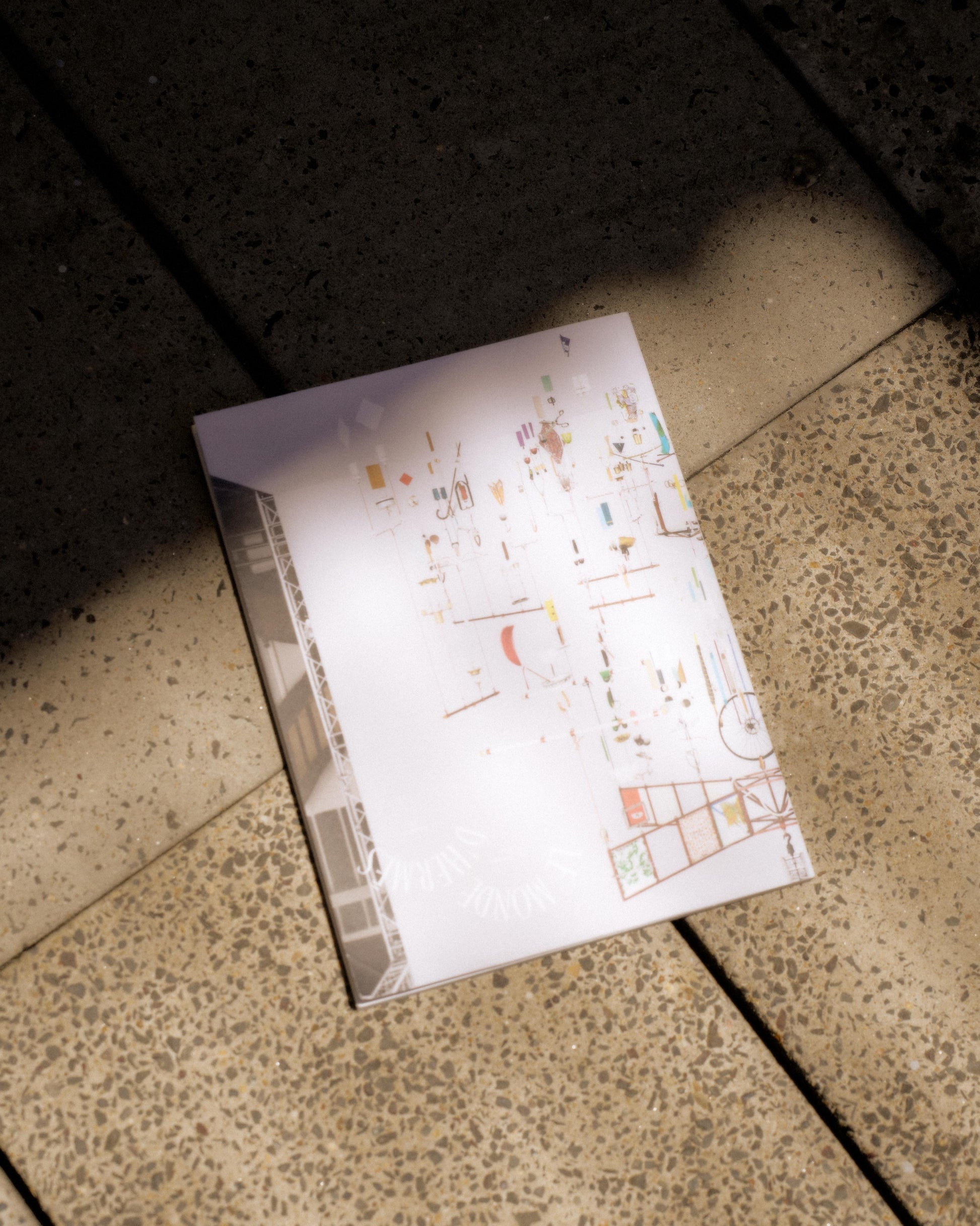 A white Hermes magazine with colourful architectural diagrams lies on a speckled stone floor, partially in shadow