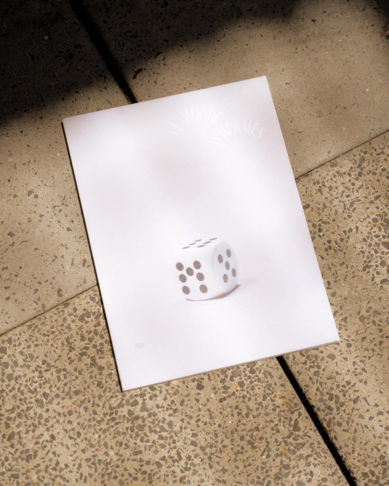 A softcover Hermes magazine with a die on the cover, placed on a textured surface.