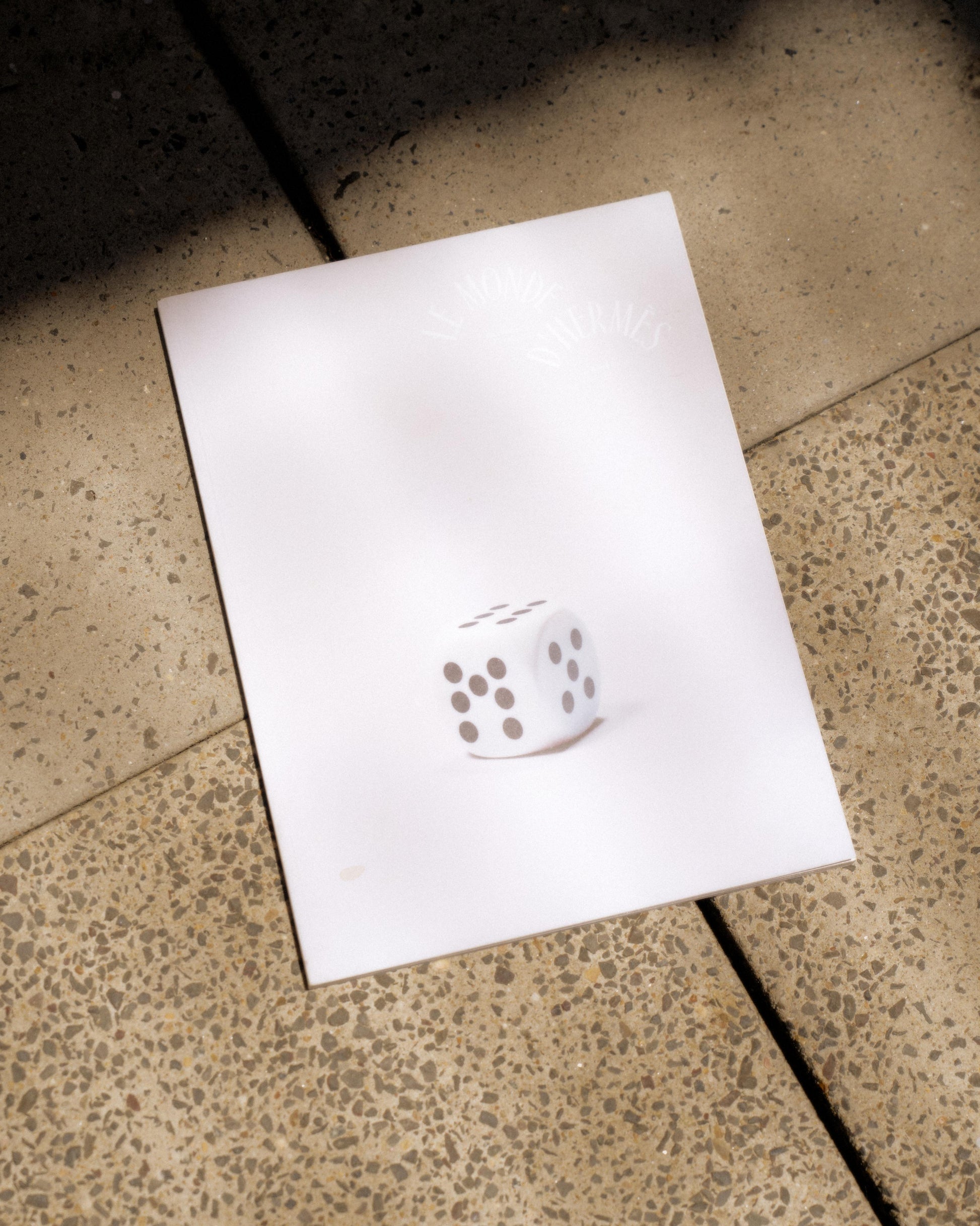 A softcover Hermes magazine with a die on the cover, placed on a textured surface.