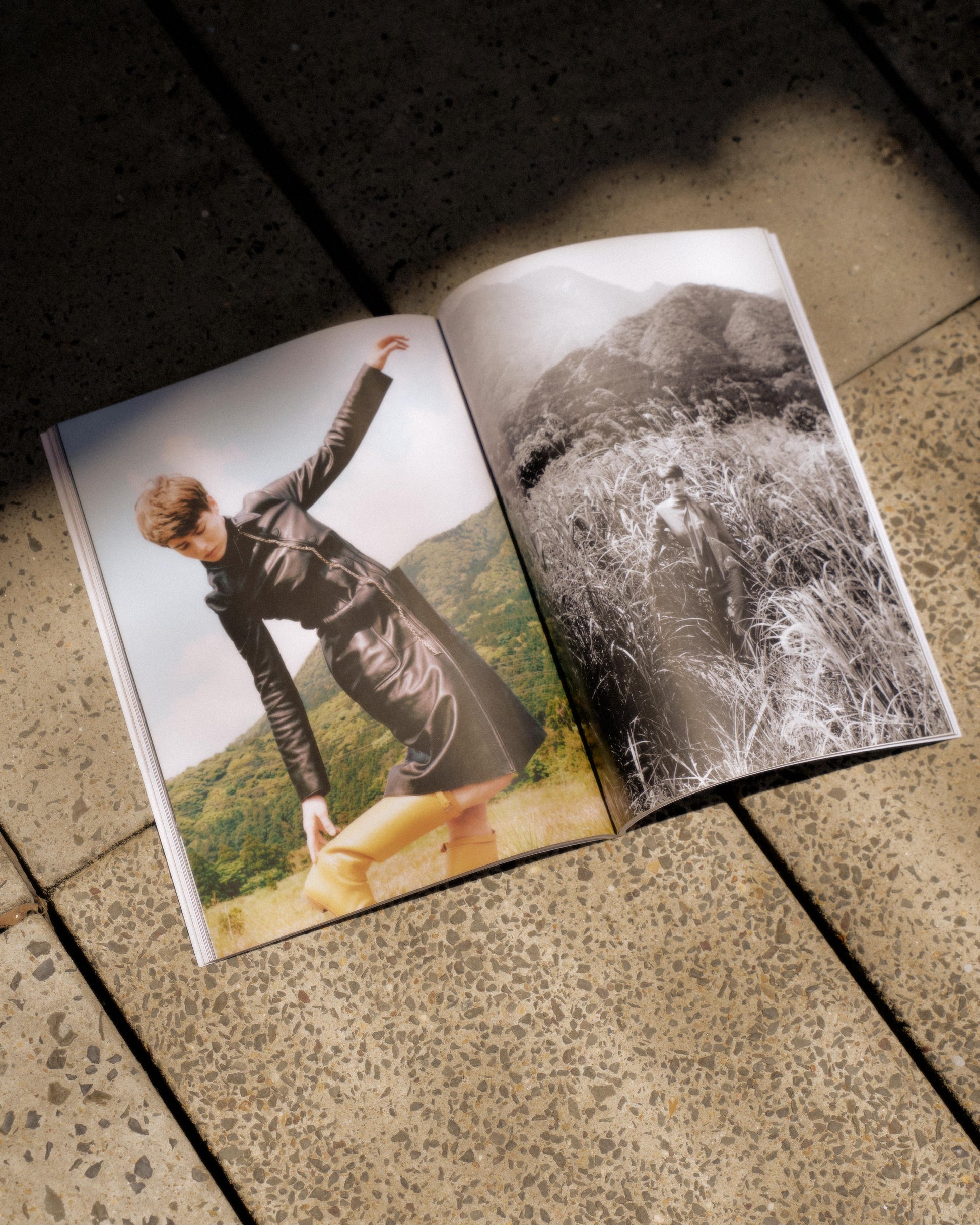 Open Hermes magazine on a tiled floor. Left page: person in a black coat leaning in a field; Right page: black-and-white image of person in tall grass with mountains.
