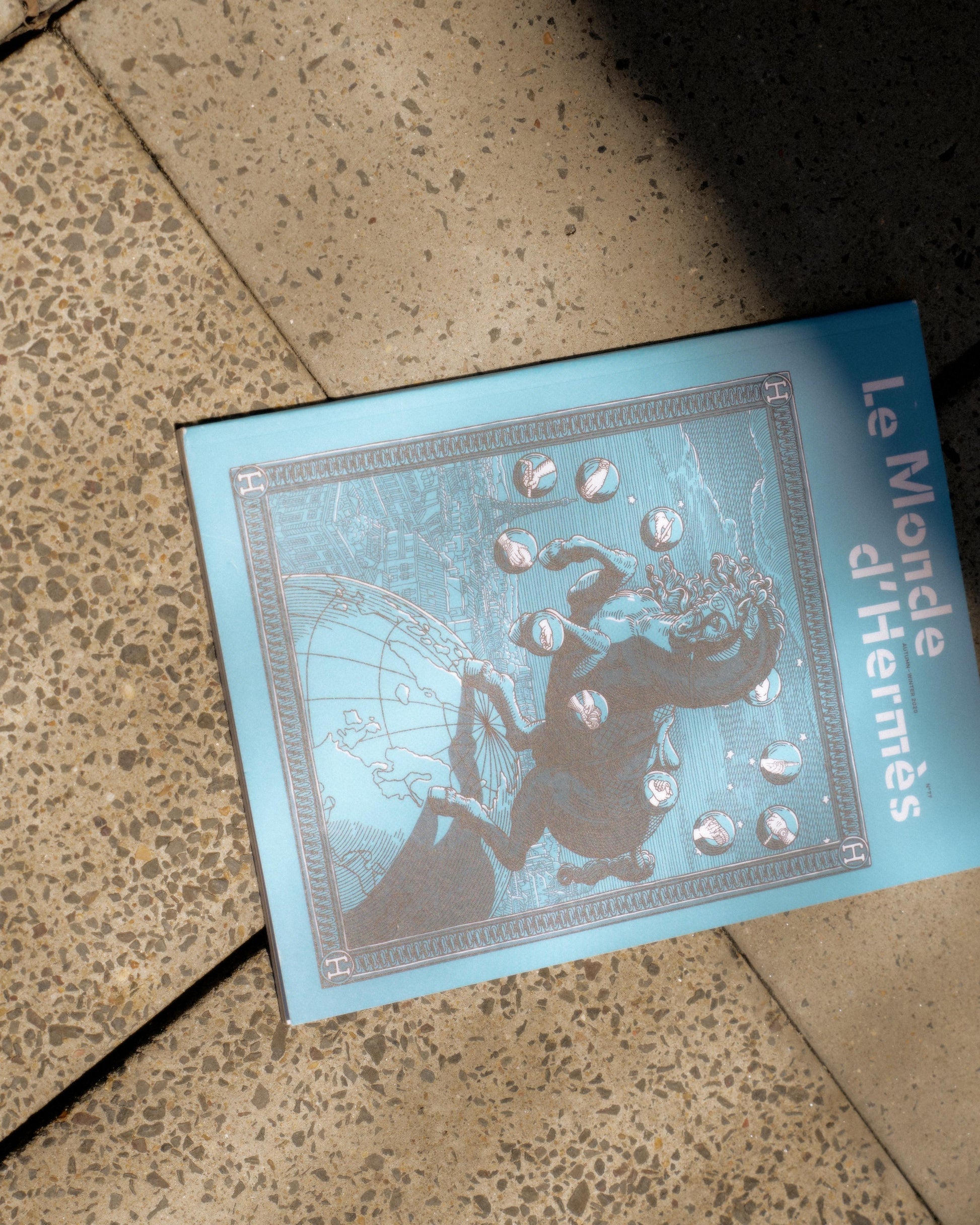Outdoor scene of "Le Monde d'Hermès" magazine with a celestial horse illustration on the cover, resting on a textured stone surface in sunlight.