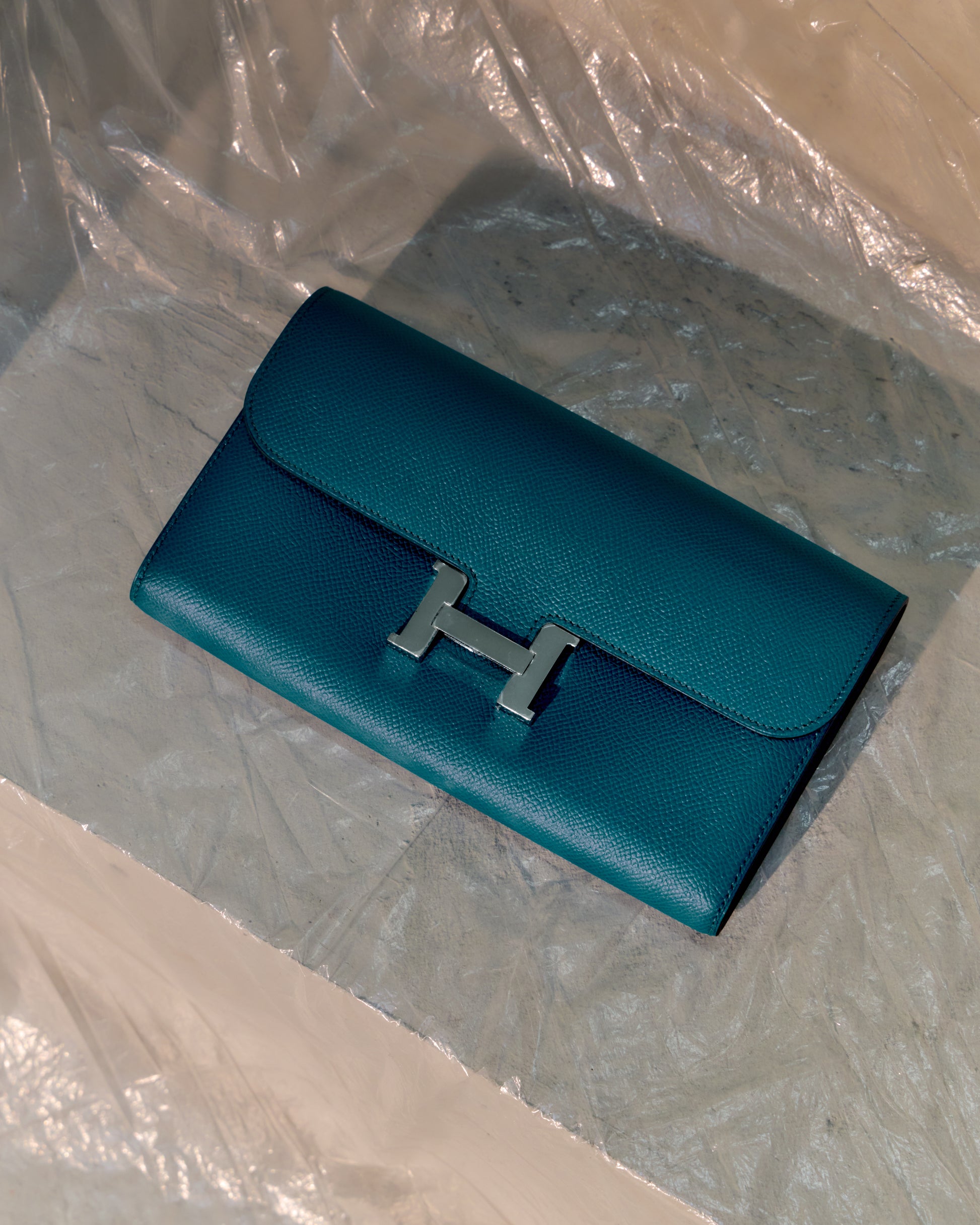 Teal Hermès wallet with silver 'H' logo on a clear plastic sheet.